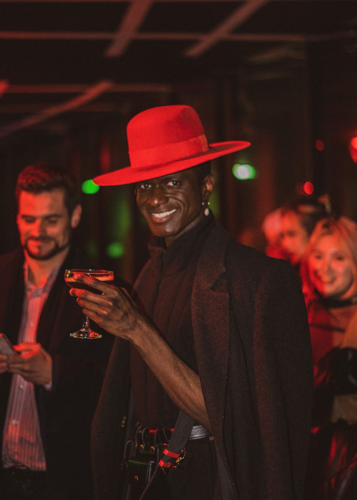 Stylish man wearing a red wide-brim hat holding a cocktail glass at a dimly lit party event.