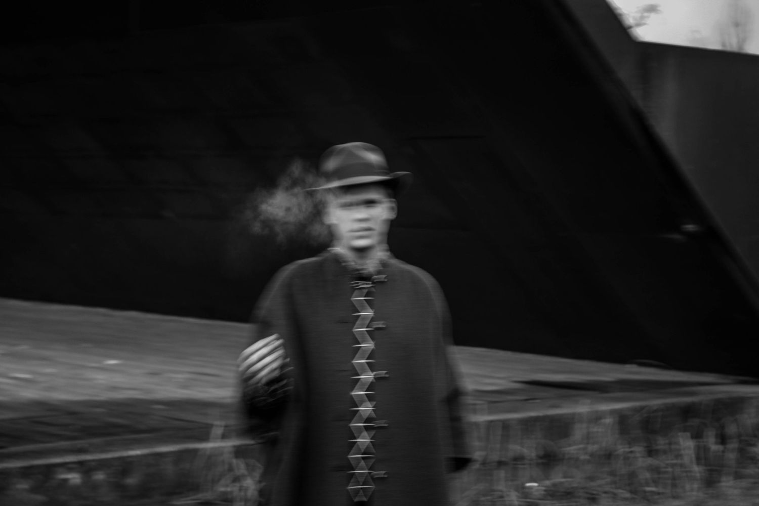 Blurry black and white photo of a person in a hat and cape smoking outdoors.