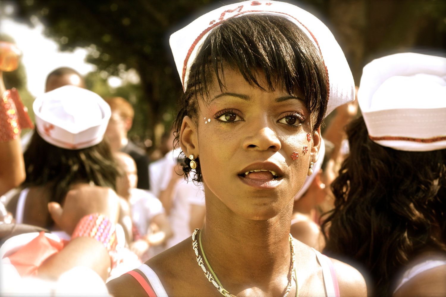 A young Black woman in a white nurse cap stands among a crowd at a vibrant outdoor cultural parade.