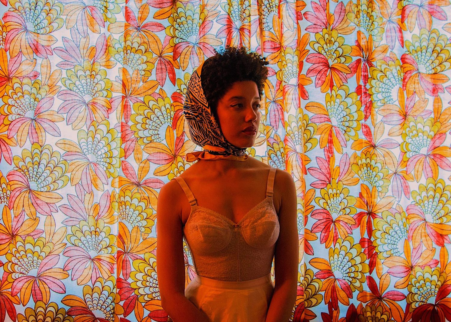 Woman in beige bralette posing before colorful floral curtain backdrop.