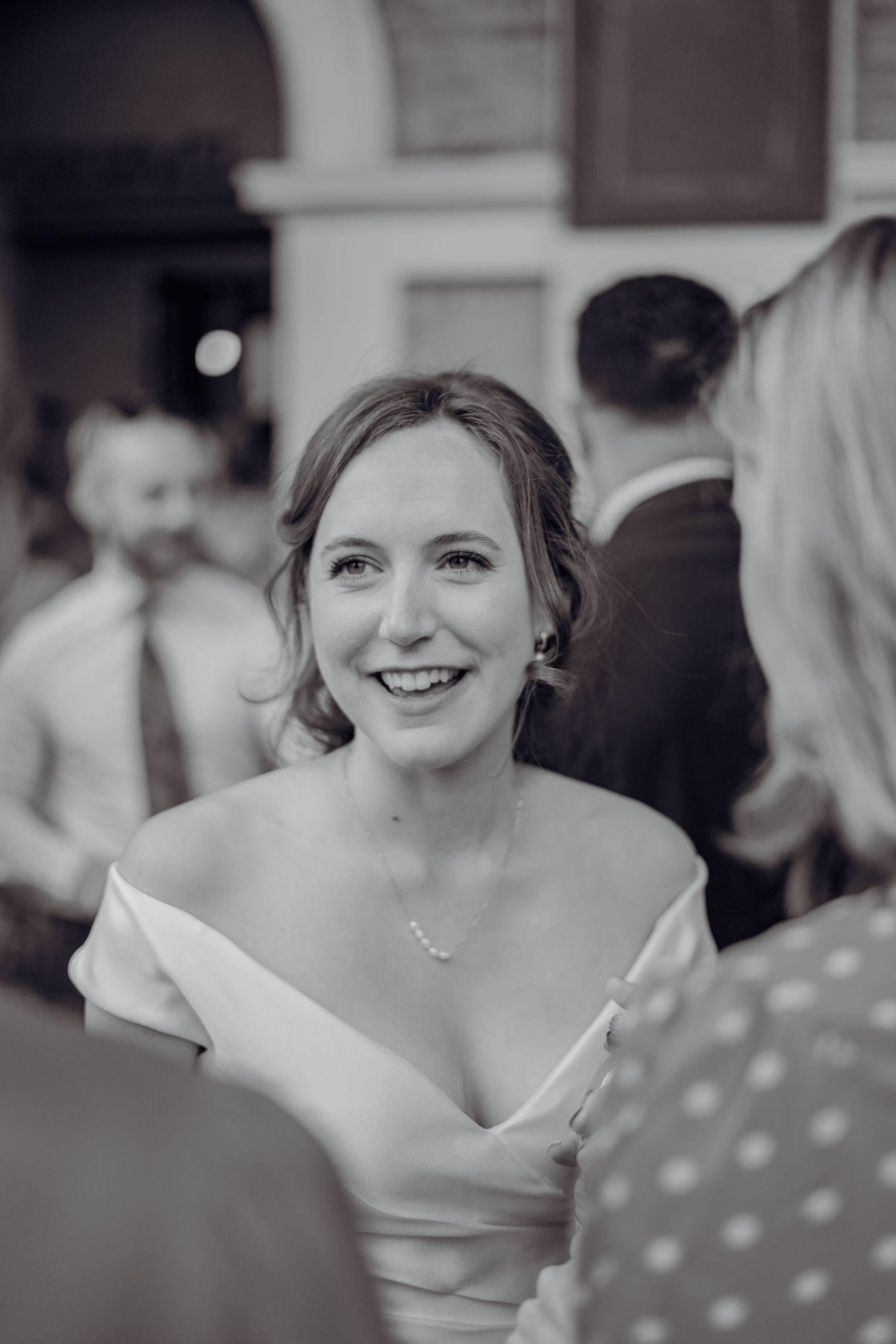 Smiling bride in off-shoulder white gown wearing pearl necklace mingling with wedding guests in black and white.