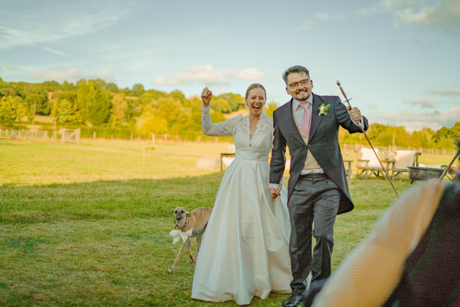 Happy bride and groom celebrating in a sunny field with a small dog following behind them.