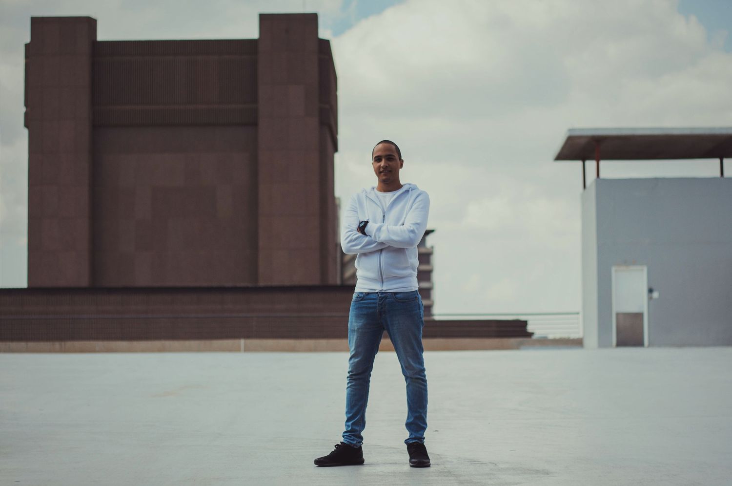 Man in white hoodie and jeans standing on rooftop with arms crossed against cloudy sky.