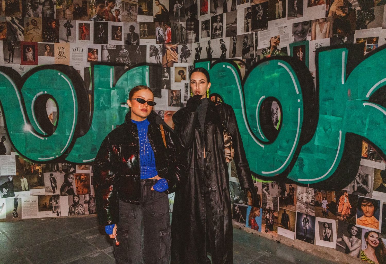 Two people posing in front of a graffiti wall covered with photos at a fashion event.