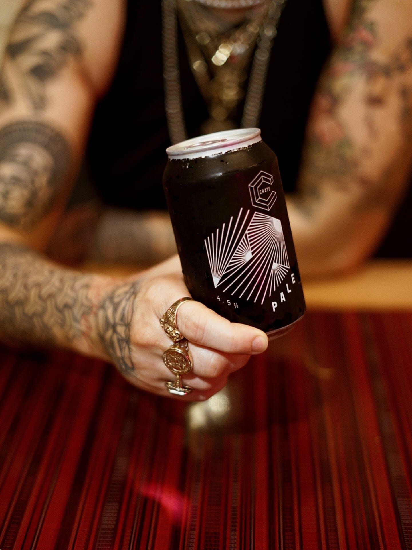 Tattooed hands holding a dark craft beer can with geometric design at a bar.
