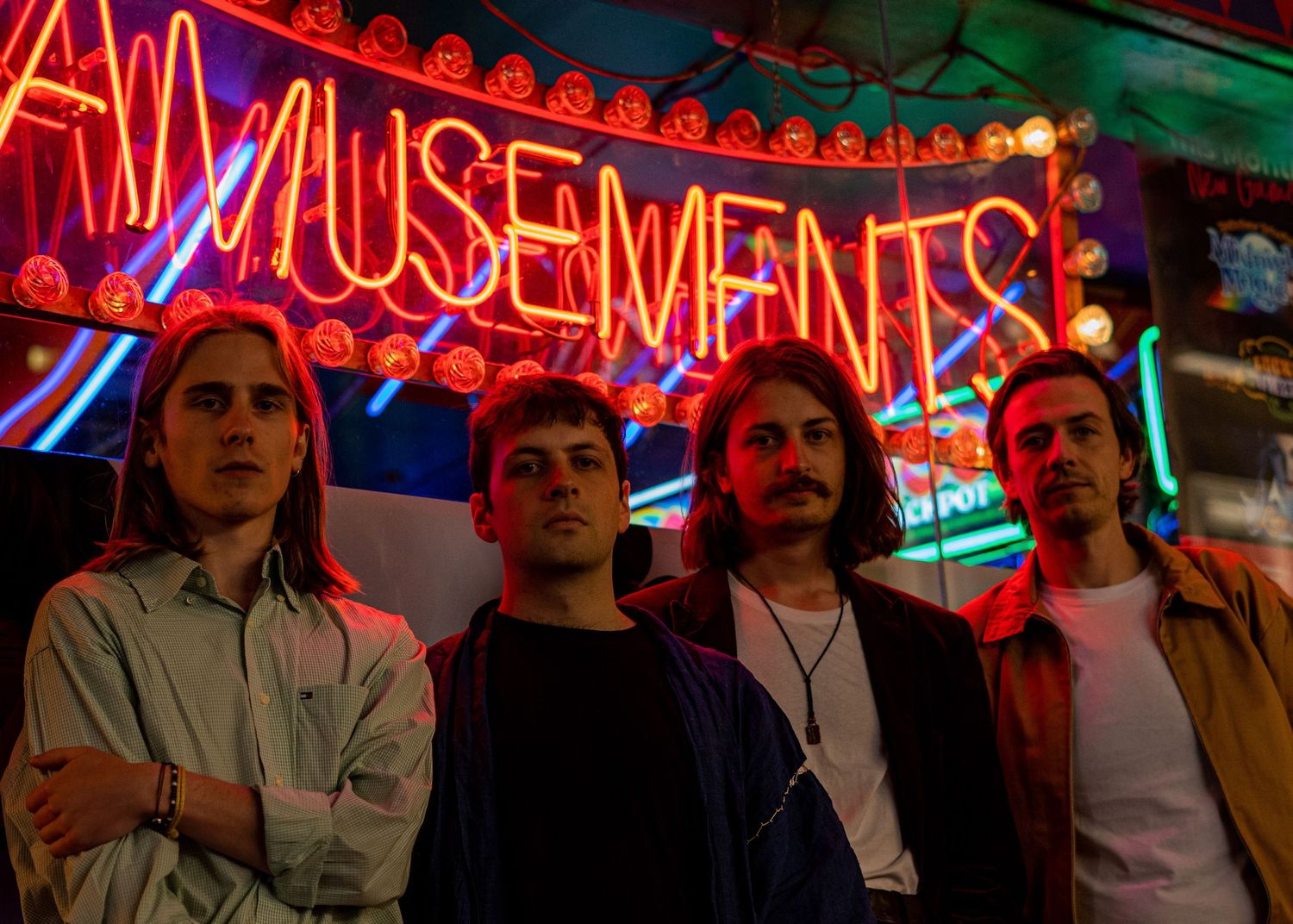 Four young men pose together in front of bright neon amusements arcade signage.