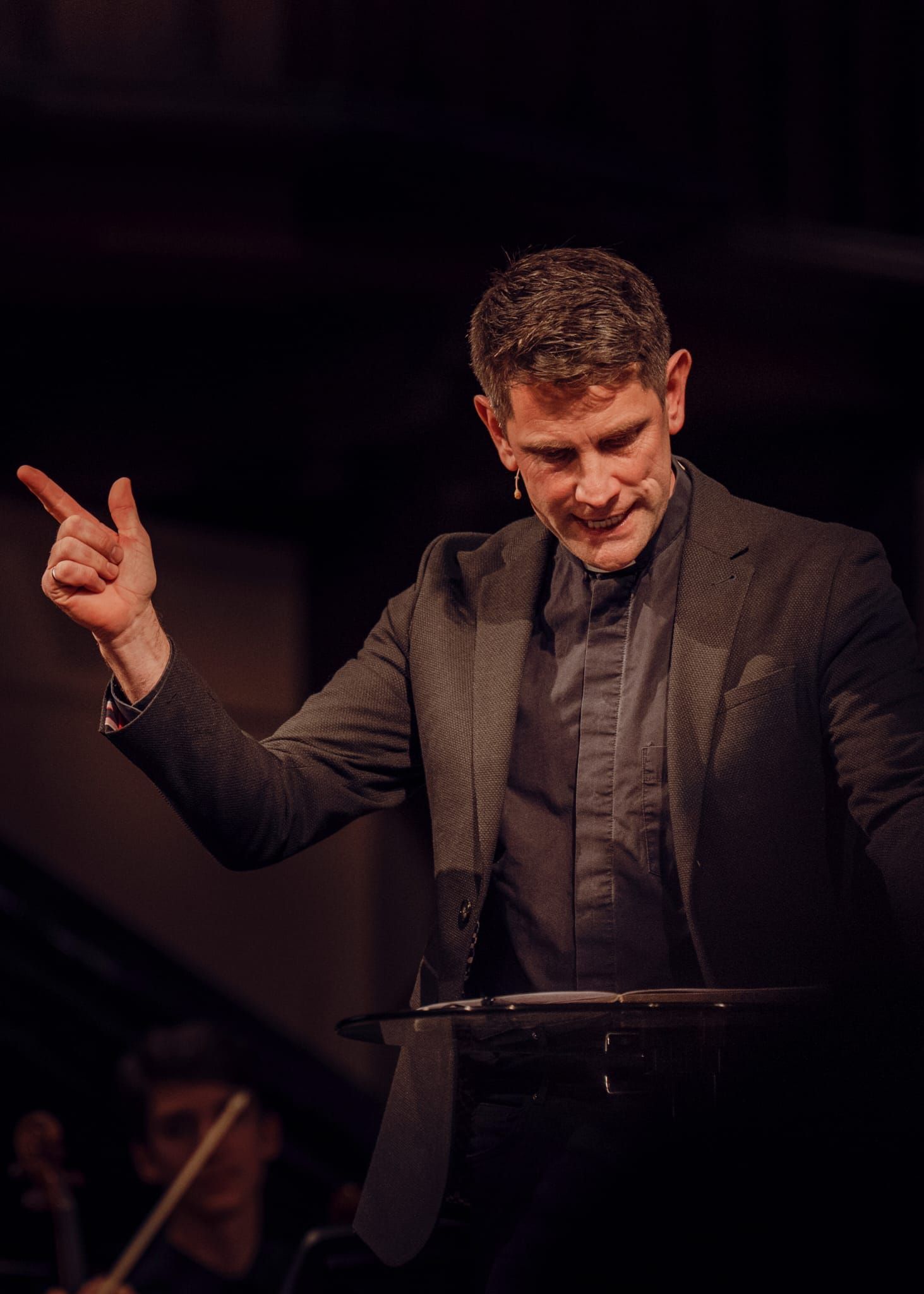 Male conductor in dark suit gesturing expressively at a classical music performance.