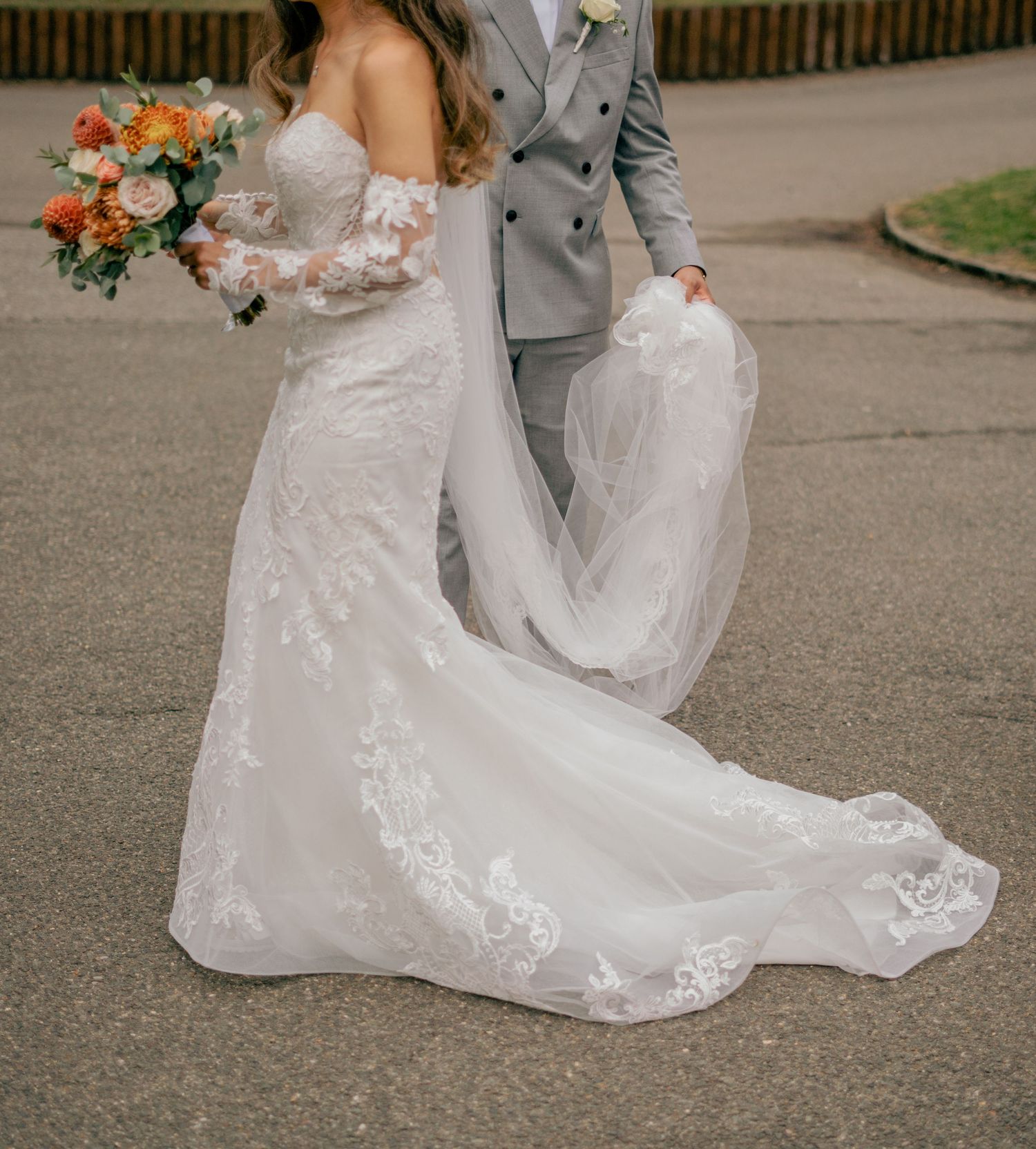 Bride in off-shoulder lace mermaid gown with floral bouquet walking with groom in gray suit.