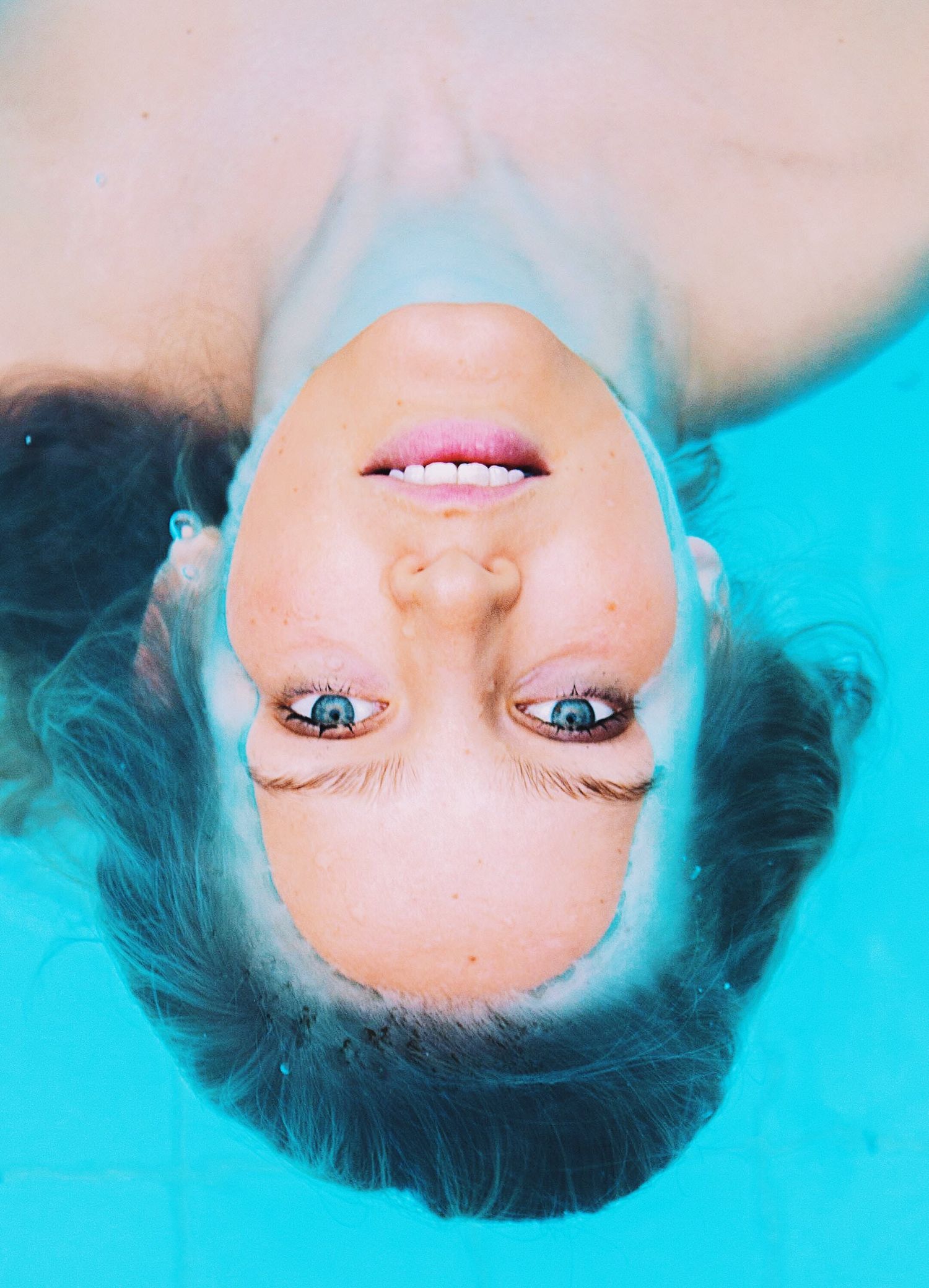 Upside-down portrait of a smiling woman with blue eyes floating in a turquoise swimming pool.
