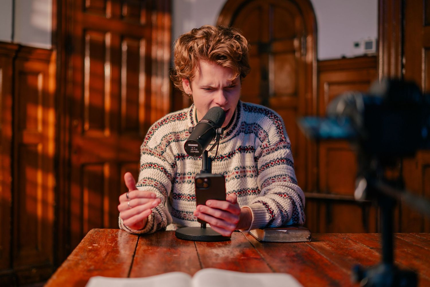 A redheaded man records a podcast using a microphone and phone at a wooden desk.
