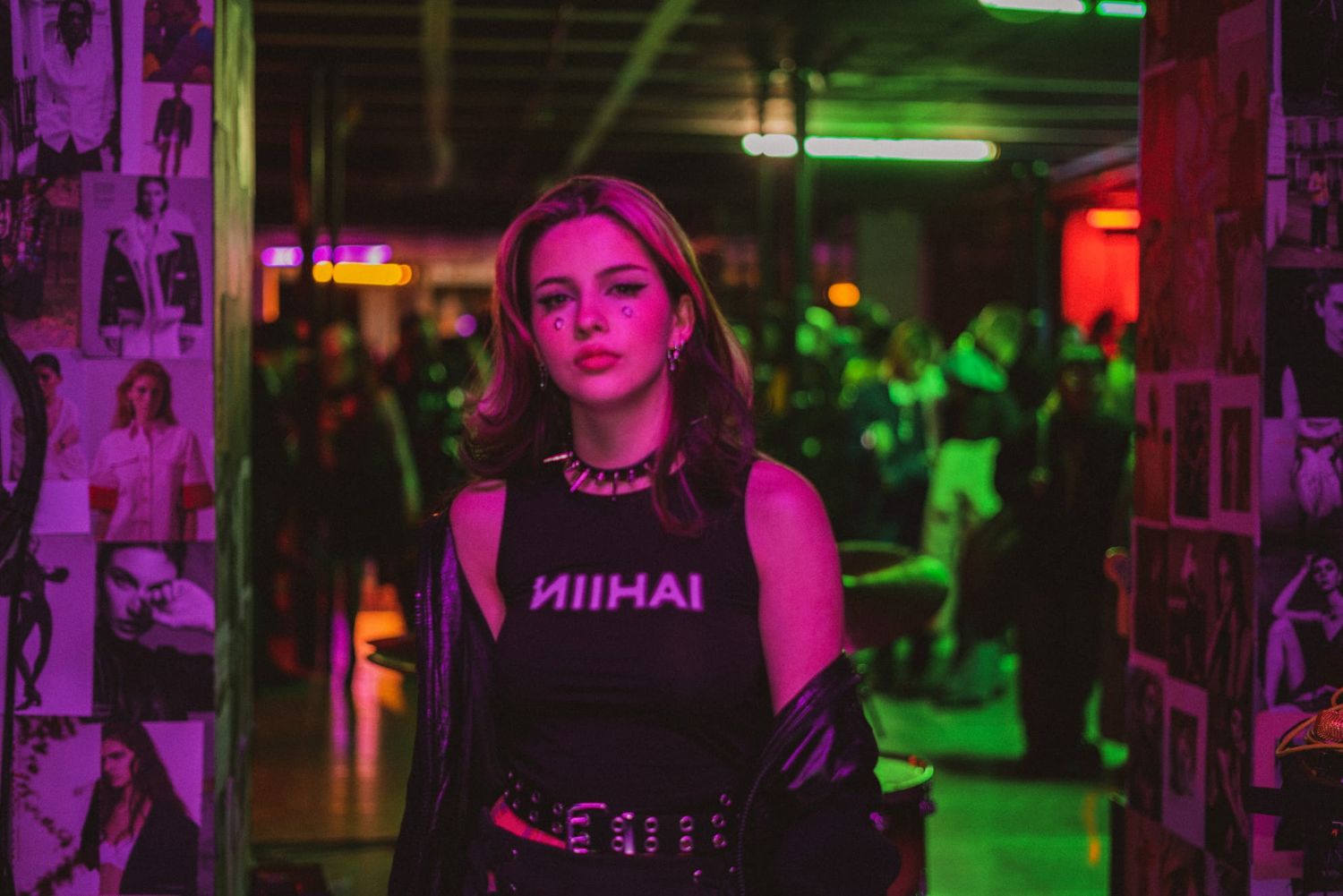 Young woman in a black graphic tee standing in a neon-lit venue with a crowd in the background.