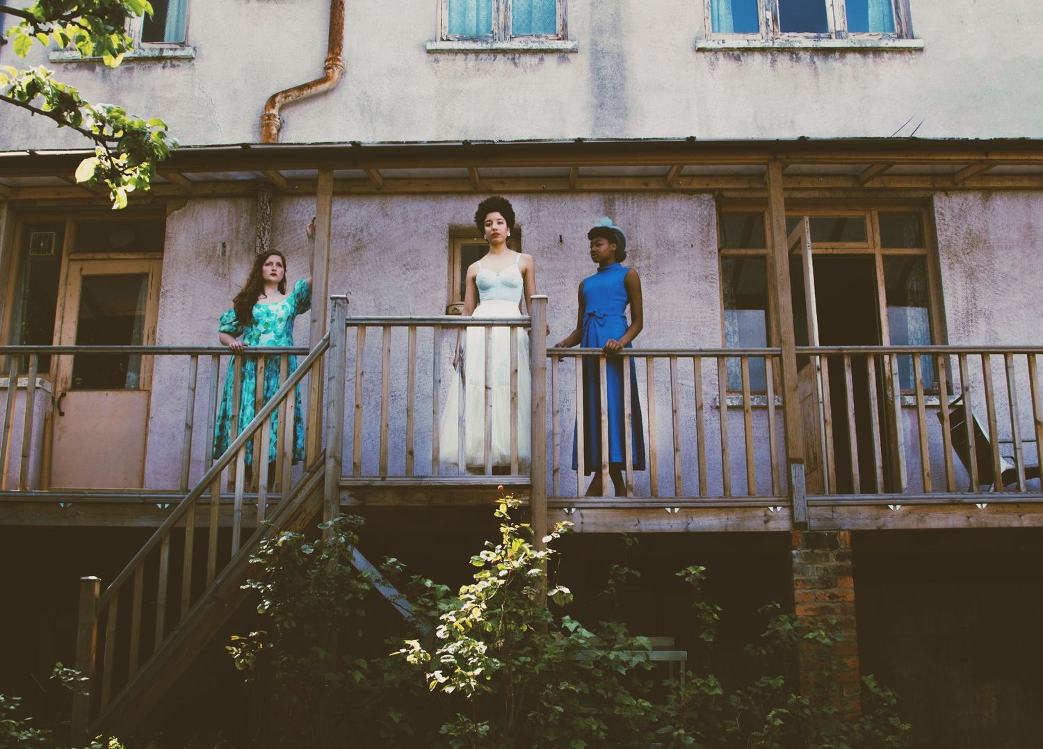Three people stand on a weathered wooden balcony of an old rustic building surrounded by trees.