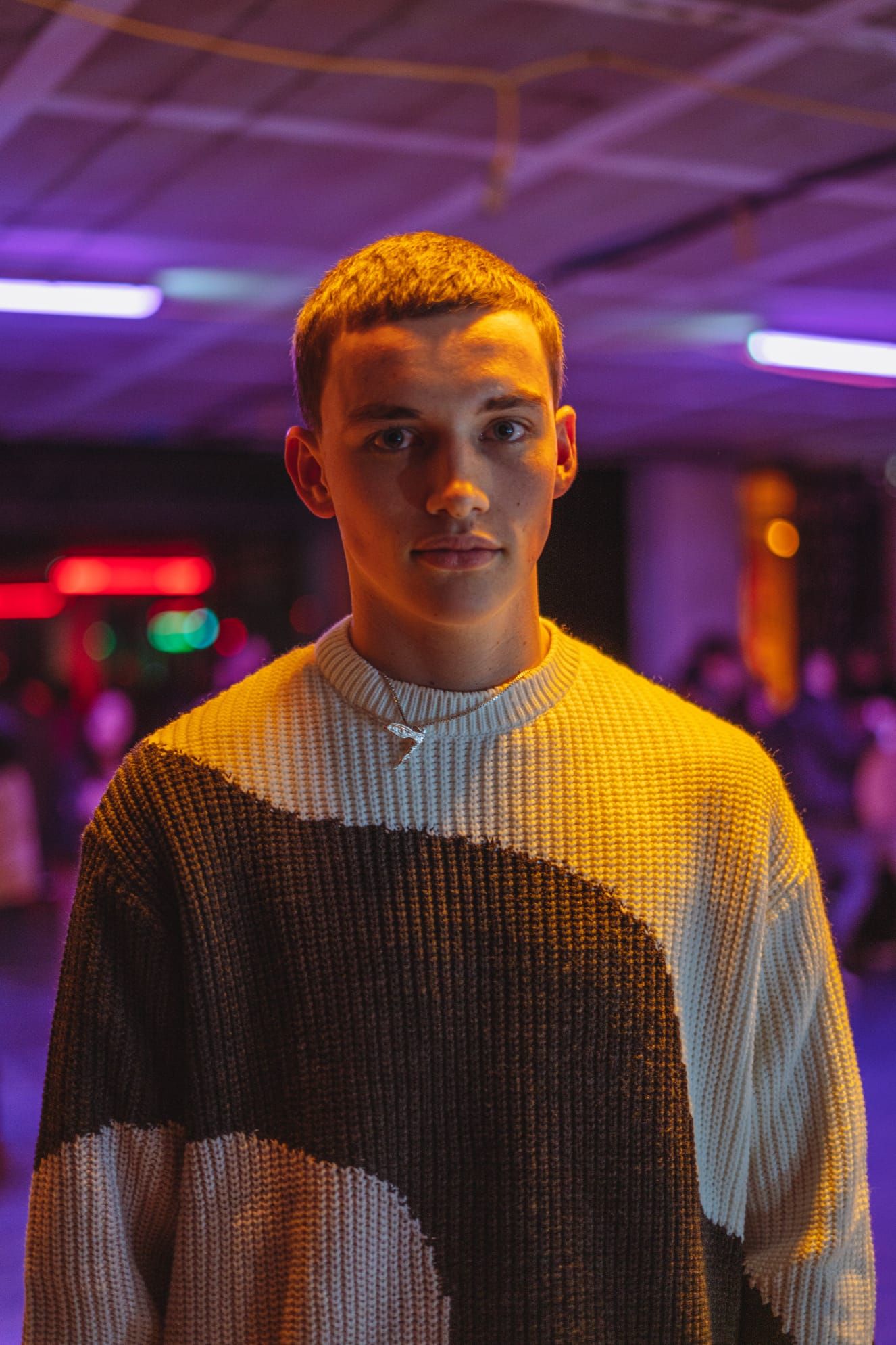 Young man in a two-tone knit sweater posing under purple neon lights at an indoor venue.