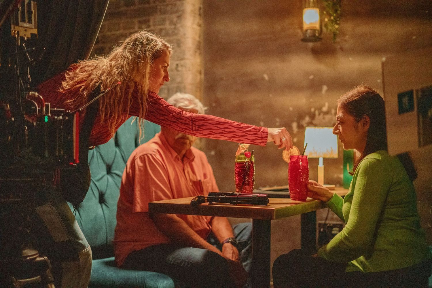 Three people sit in a cozy bar booth as a woman with red hair reaches across the table.