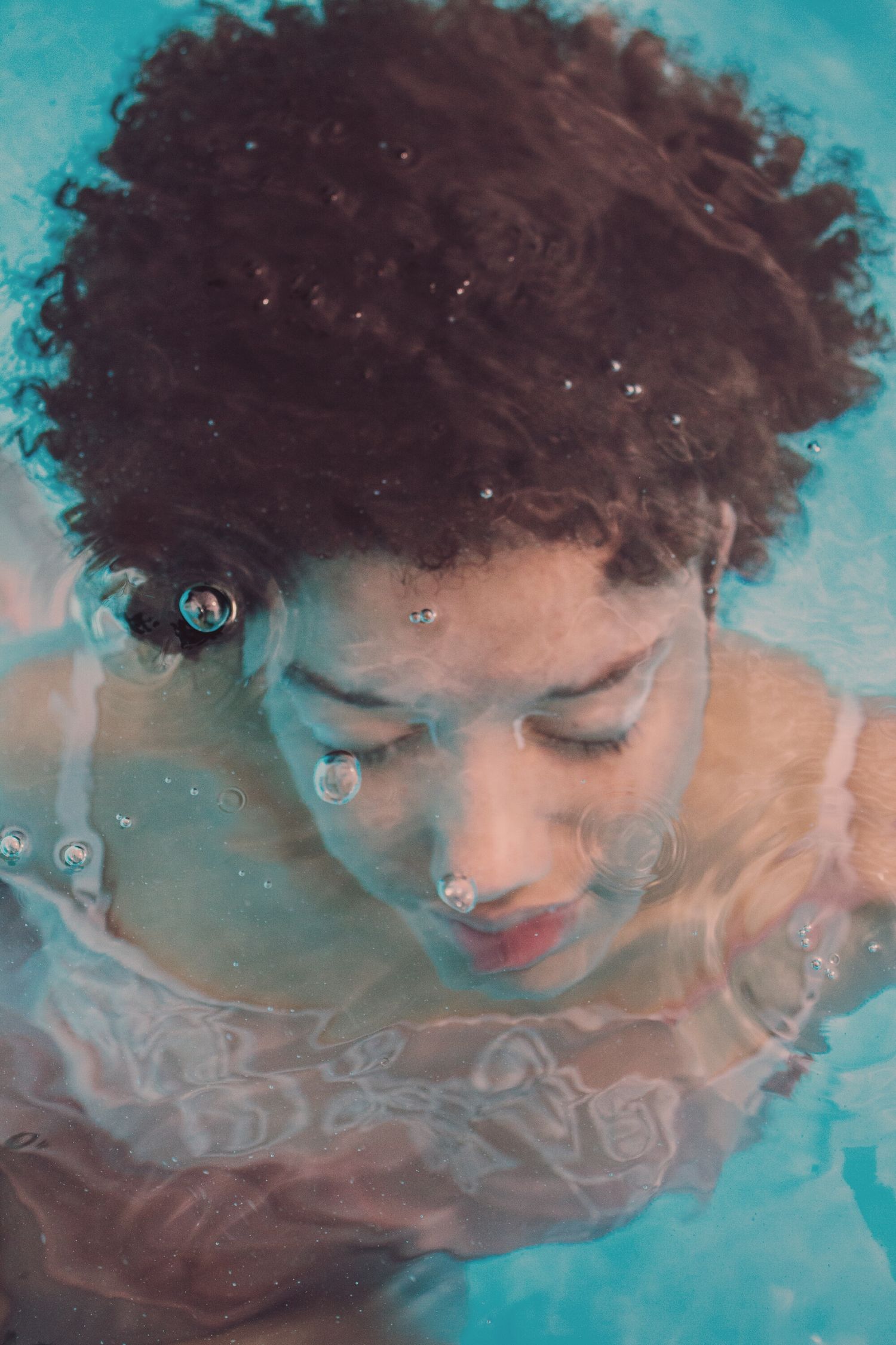 A young woman with curly afro hair submerged underwater in a blue swimming pool with bubbles.