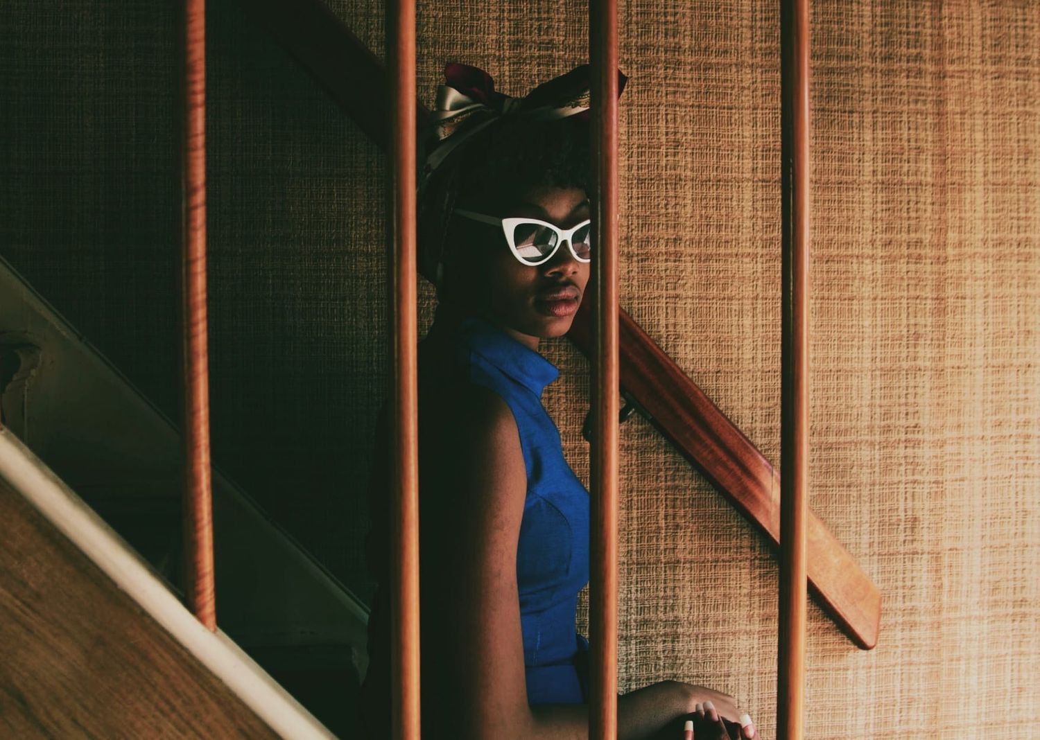 Woman in blue dress with cat ear headband and white cat-eye glasses peering through stair railings.