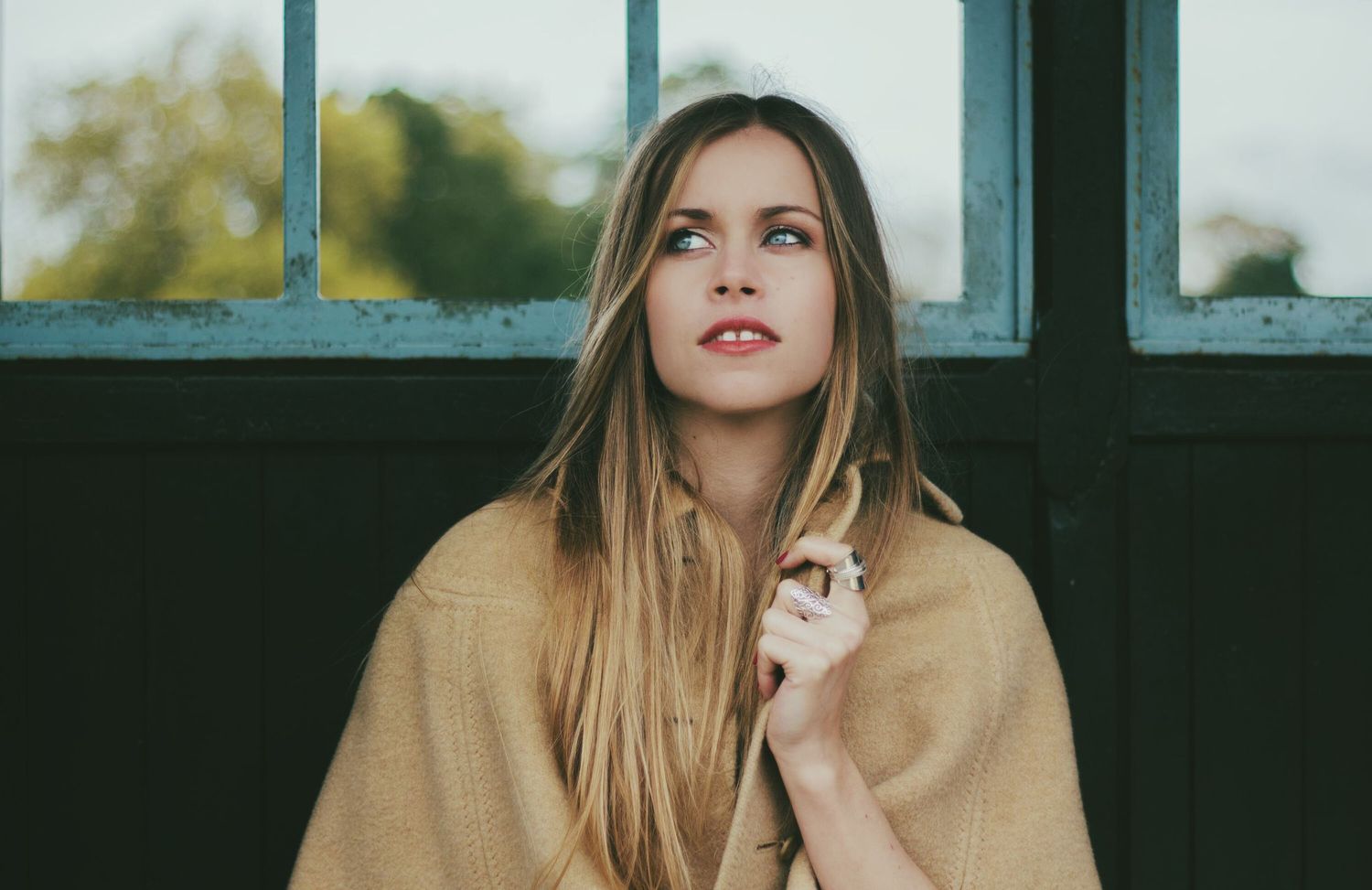 A blonde woman wrapped in a tan blanket gazes upward near dark industrial windows in soft light.