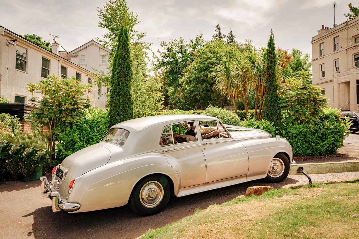 Vintage silver Rolls Royce wedding car parked outside an elegant building surrounded by tall trees.
