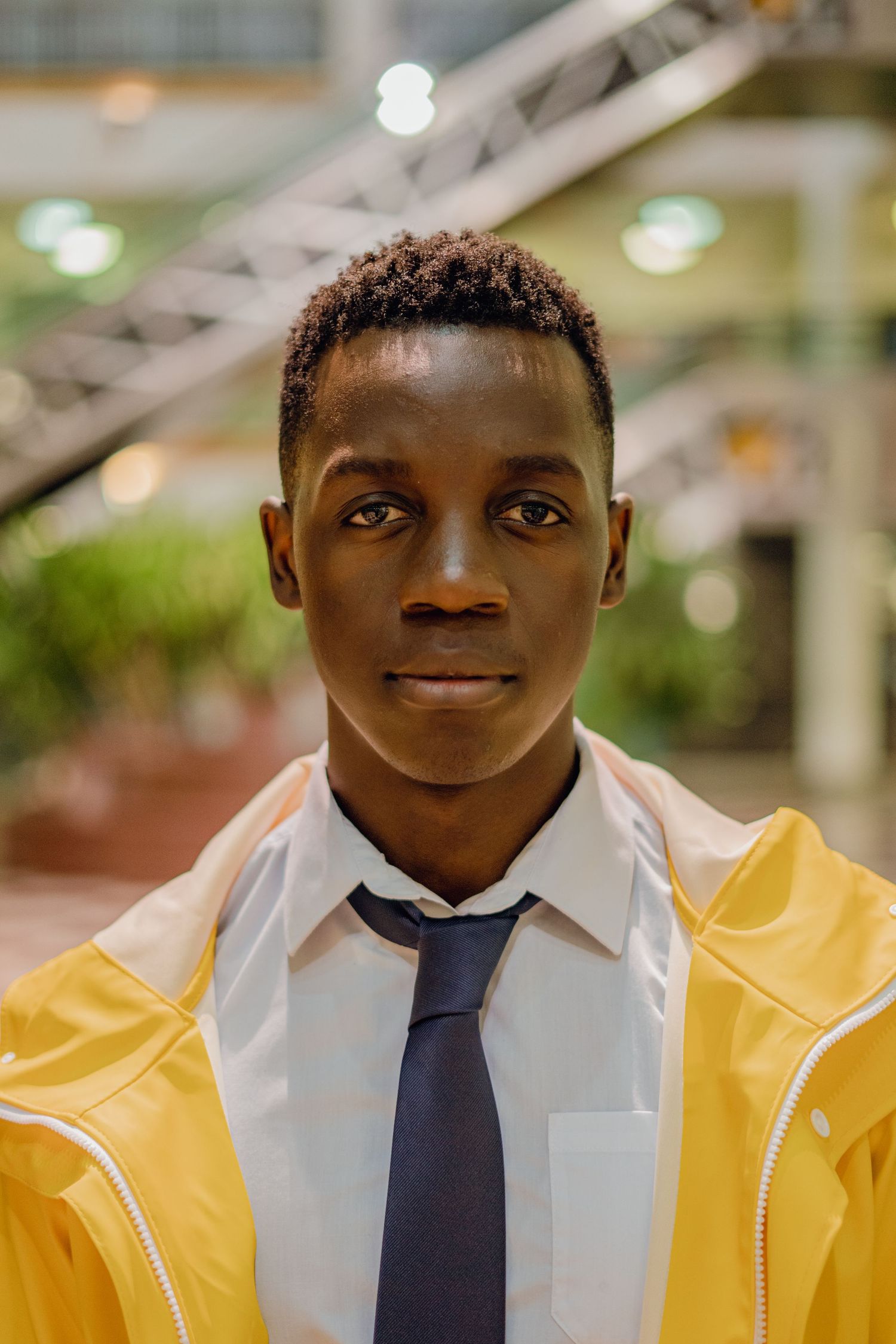 Young African man in yellow jacket and tie poses confidently indoors under bright lights.