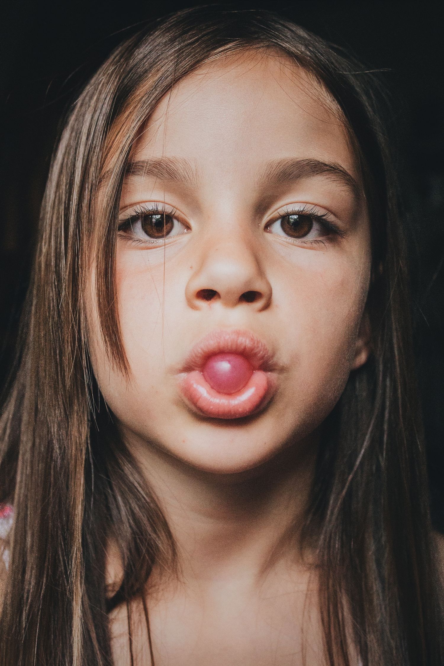 A young girl with long brown hair blows a pink bubble gum bubble against a dark background.