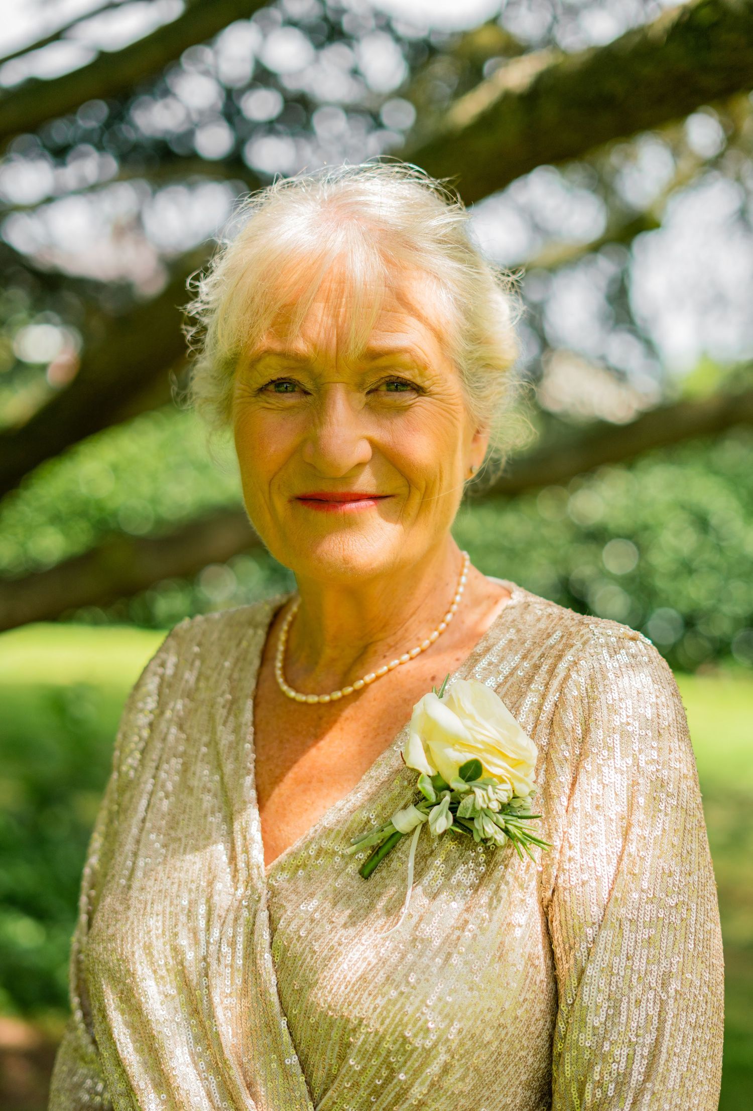 Elegant older woman in gold sequin dress with white floral corsage and pearl necklace smiling at a wedding.