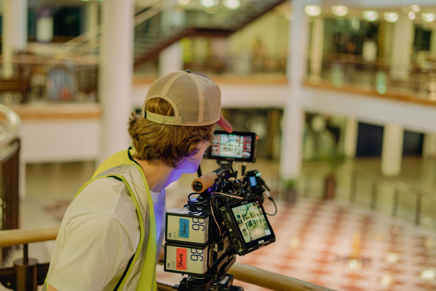 A cameraman films with a professional camera rig inside a grand building with balconies.