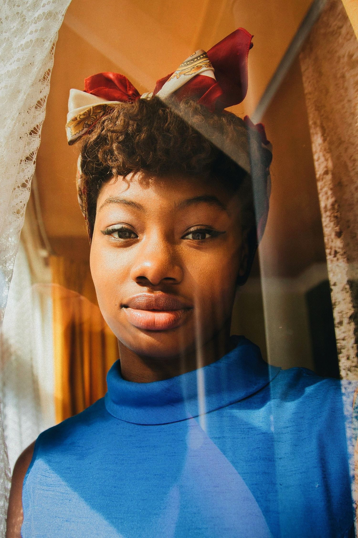 Young woman in blue turtleneck with red bow headband photographed through glass window.