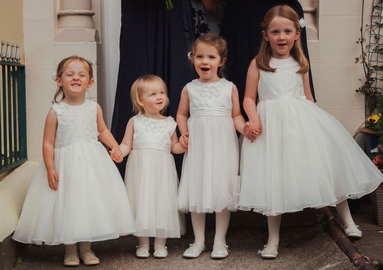 Four young flower girls in white dresses holding hands and smiling at a wedding ceremony.