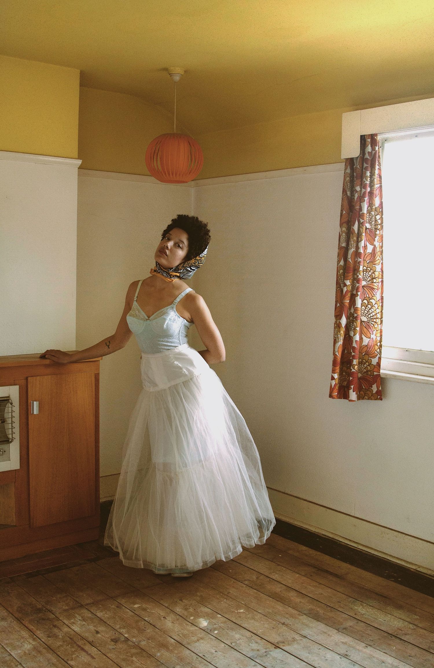 A woman in a vintage light blue tulle ball gown poses in a retro-styled room with wooden floors.
