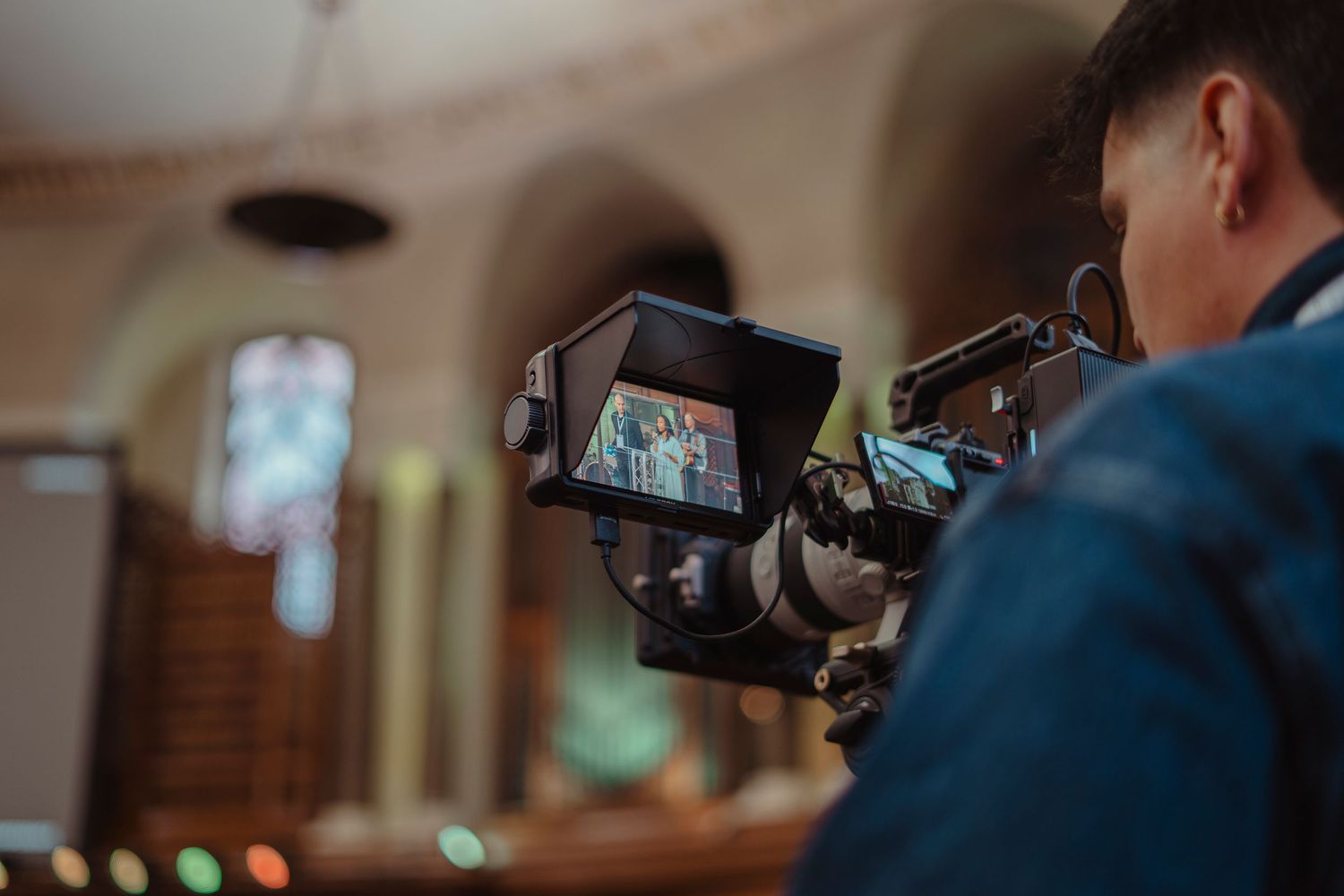 Videographer filming a wedding ceremony inside a church with stained glass windows using a professional camera.