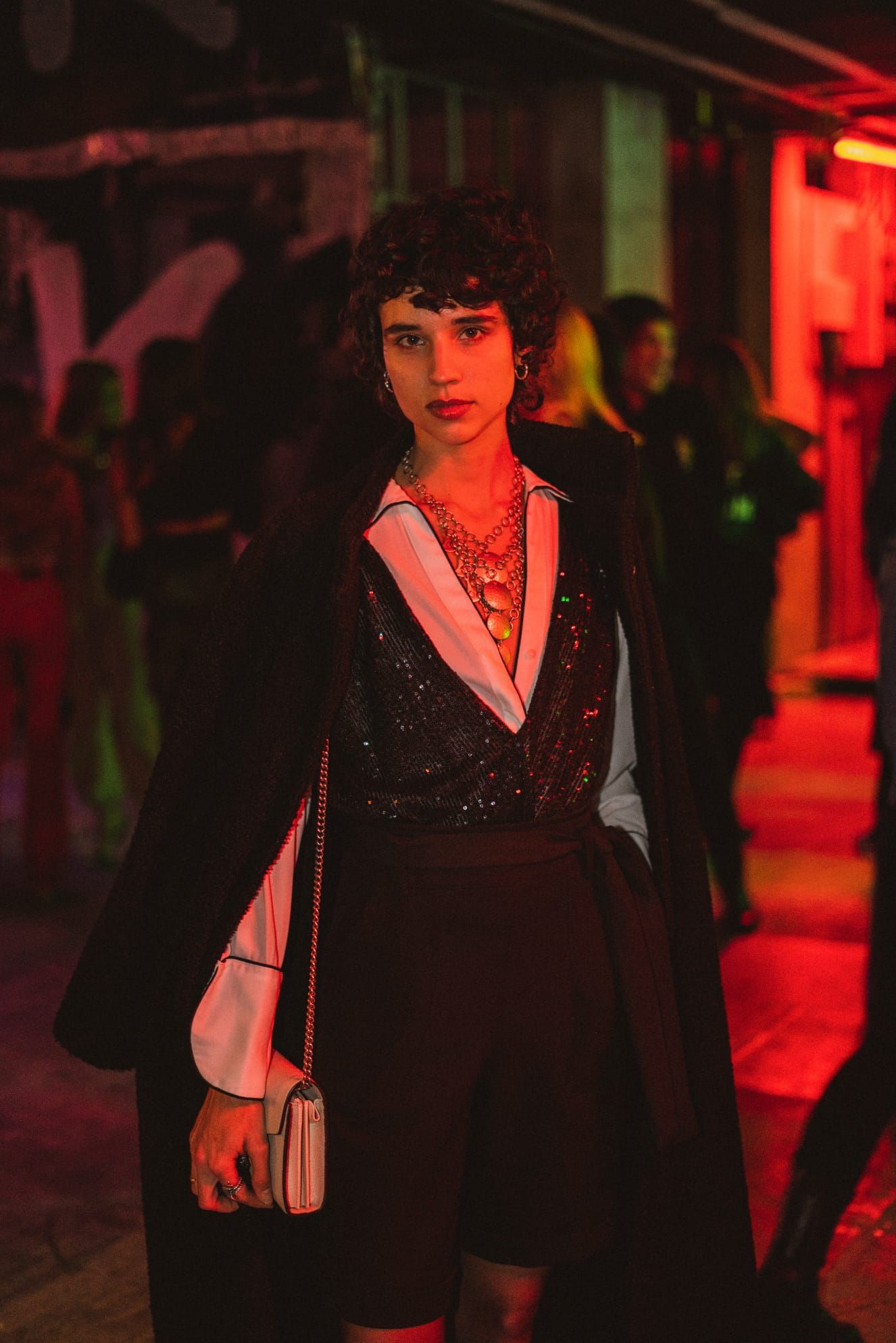 Person in sequined top and black cape coat poses under red neon lights at night event.