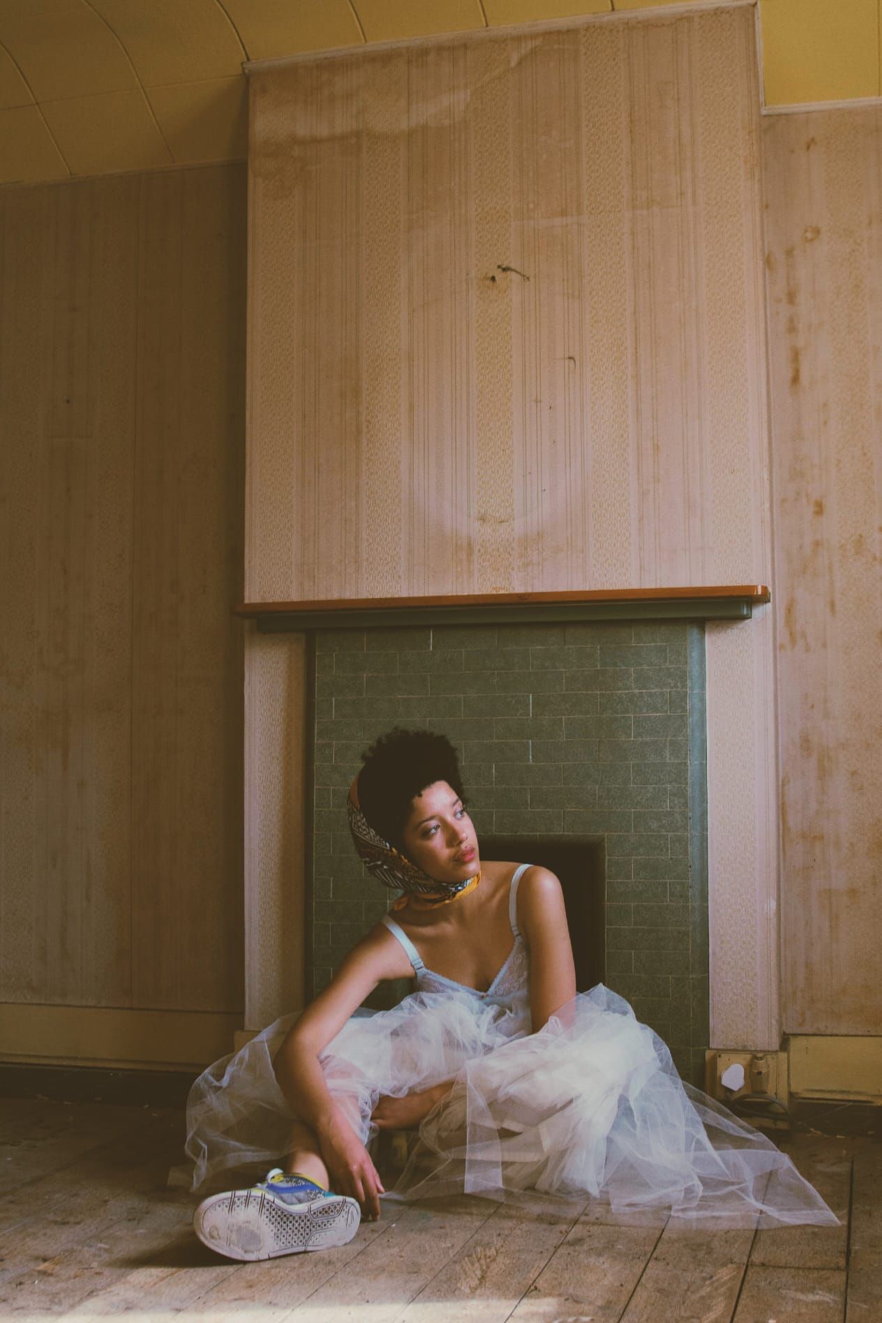 Woman in white tulle dress sitting on wooden floor in front of an old fireplace indoors.