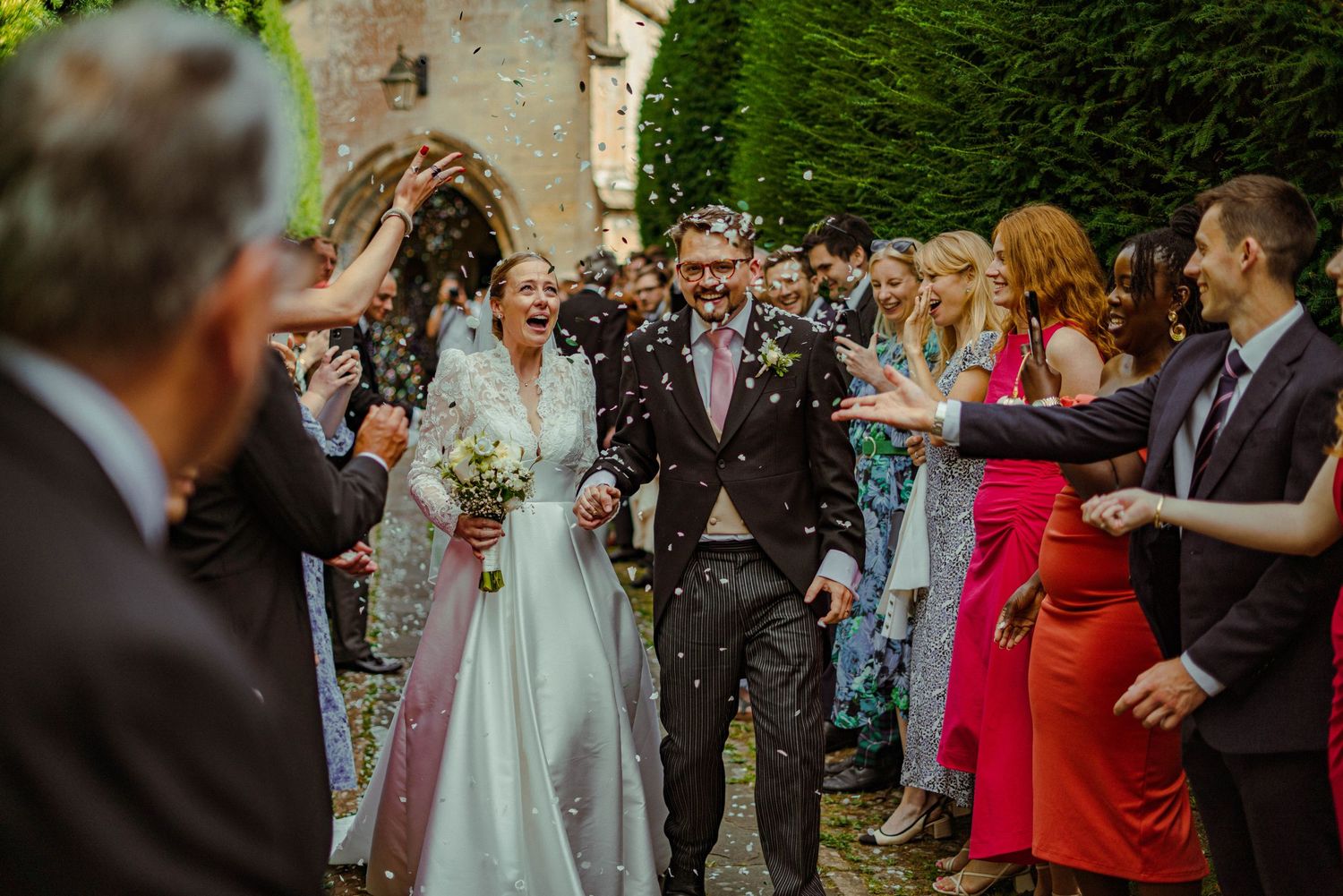 Joyful newlyweds walking through confetti shower surrounded by cheering guests outside a historic stone church.