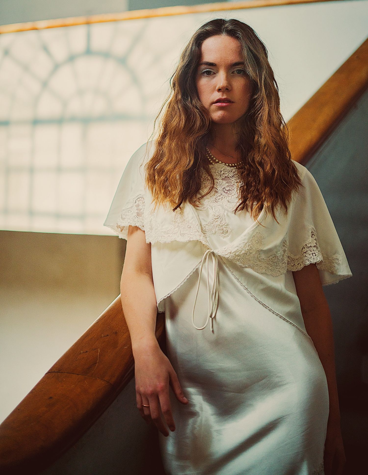 A woman in a white lace dress poses gracefully on a staircase by a arched window.