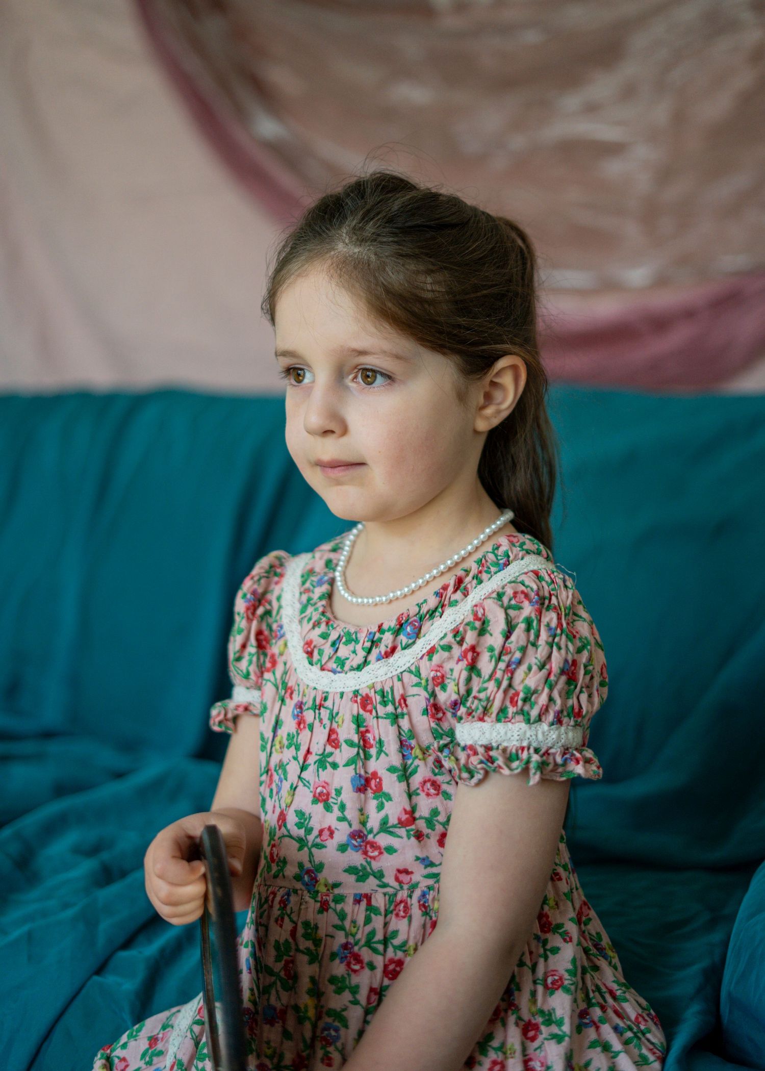 A young girl in a floral dress with a pearl necklace sits on a teal sofa holding a cane.