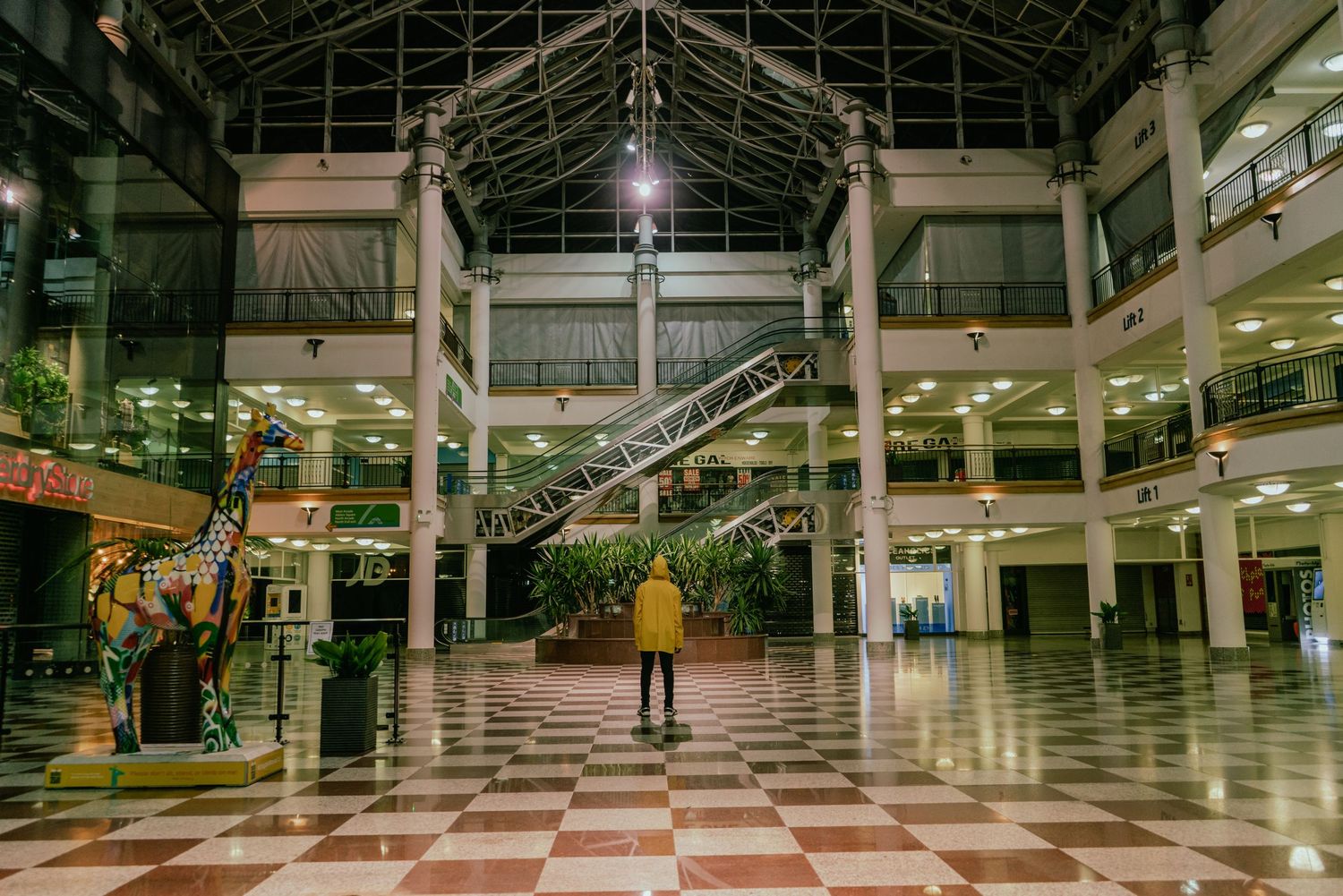 A lone person stands in a vast empty shopping mall atrium with checkered floors.