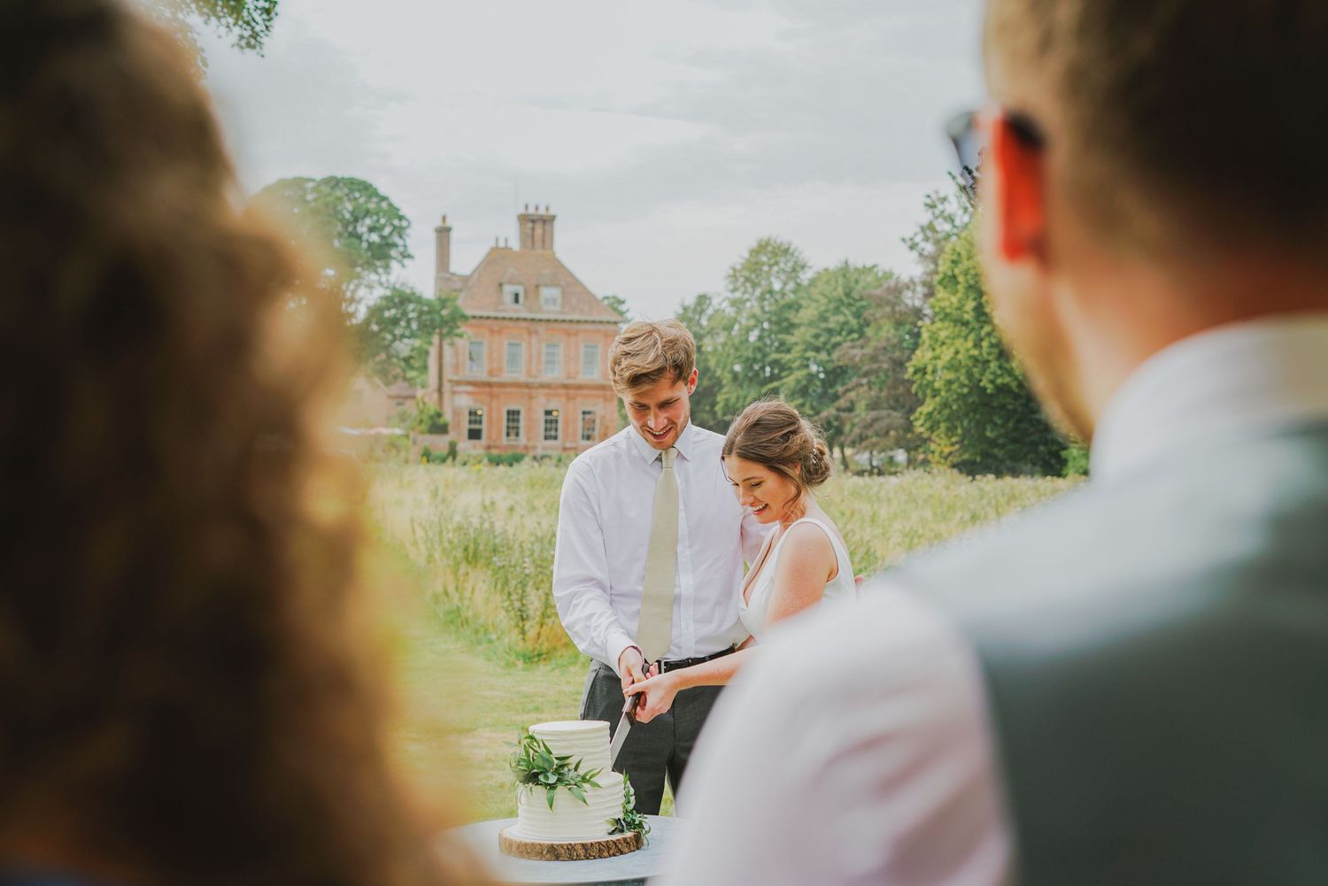 Bride and groom cutting their wedding cake outdoors with a grand country house in the background.