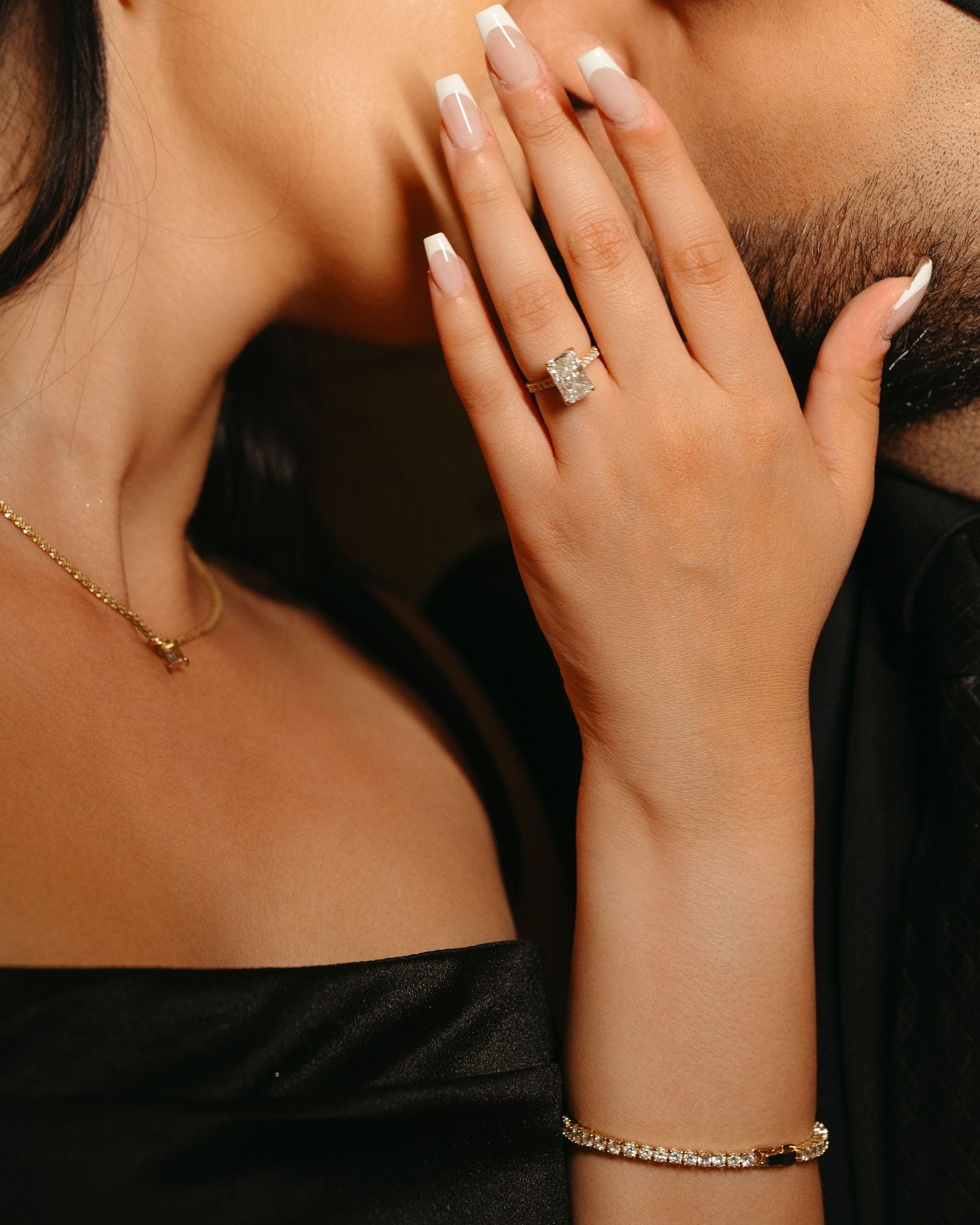 Close-up of a woman's hand with a French manicure and sparkling diamond engagement ring touching a bearded man's face.