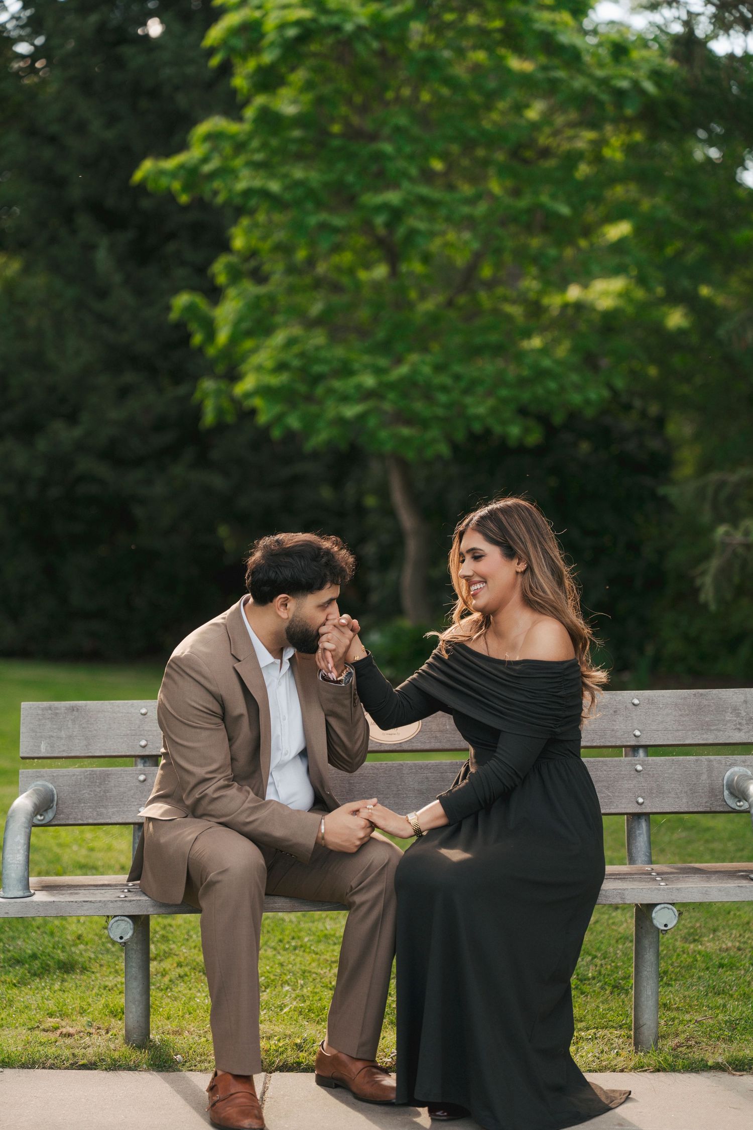 Man in a tan suit kisses his fiancée's hand as she smiles on a park bench surrounded by greenery.
