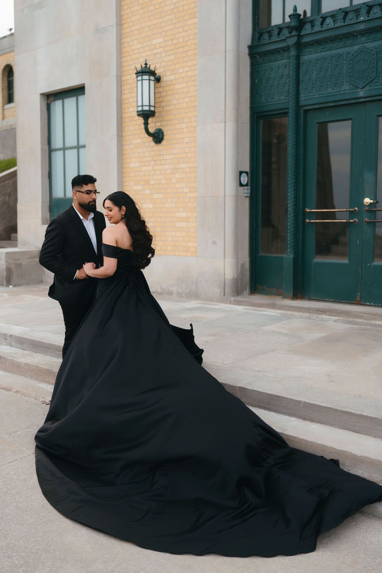 Bride in an off-shoulder black wedding gown with a dramatic train stands beside her groom in a black suit outside a green-doored building.