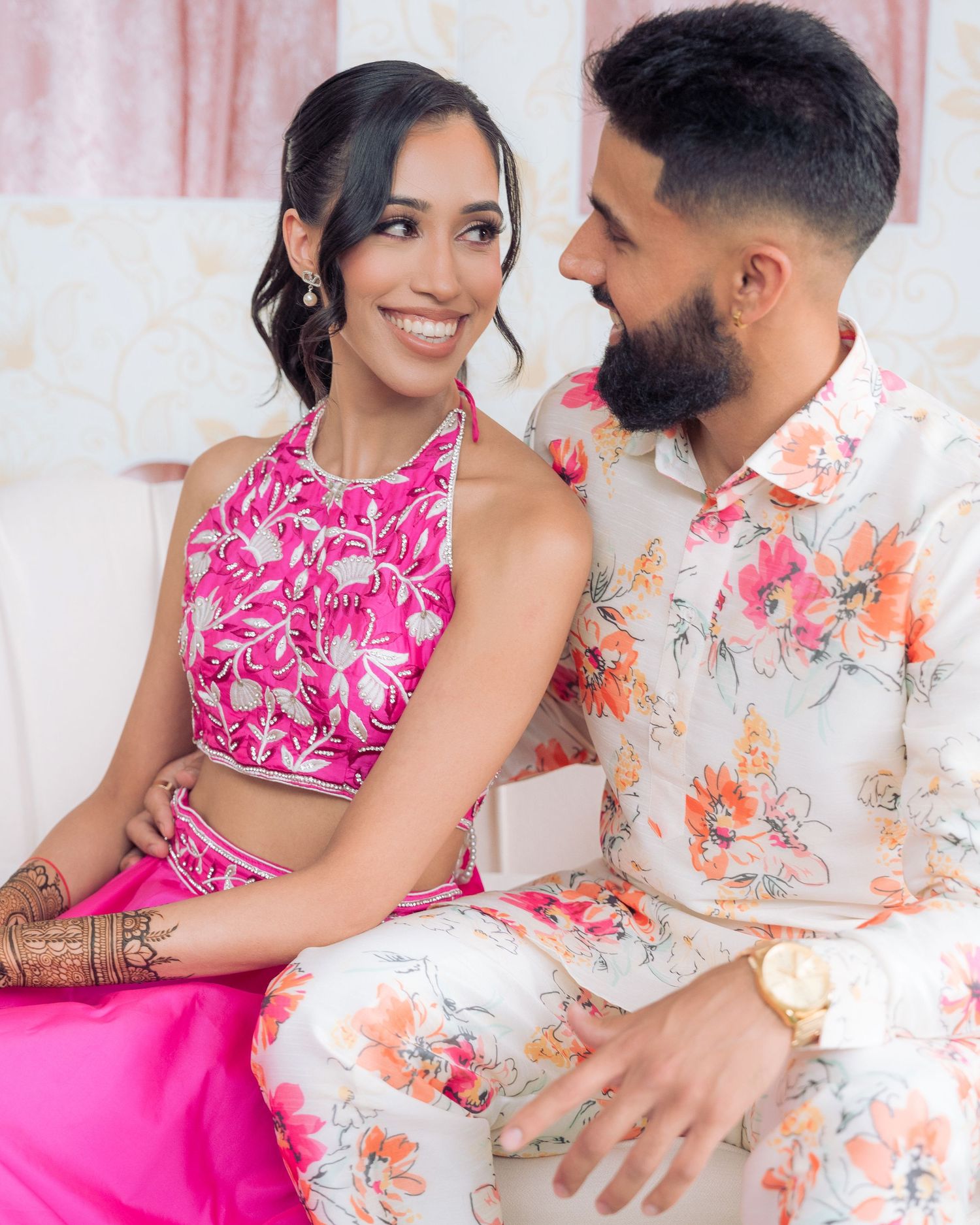 Smiling woman in a pink embroidered halter top leans into a bearded man in a floral shirt during a joyful couple's photoshoot.
