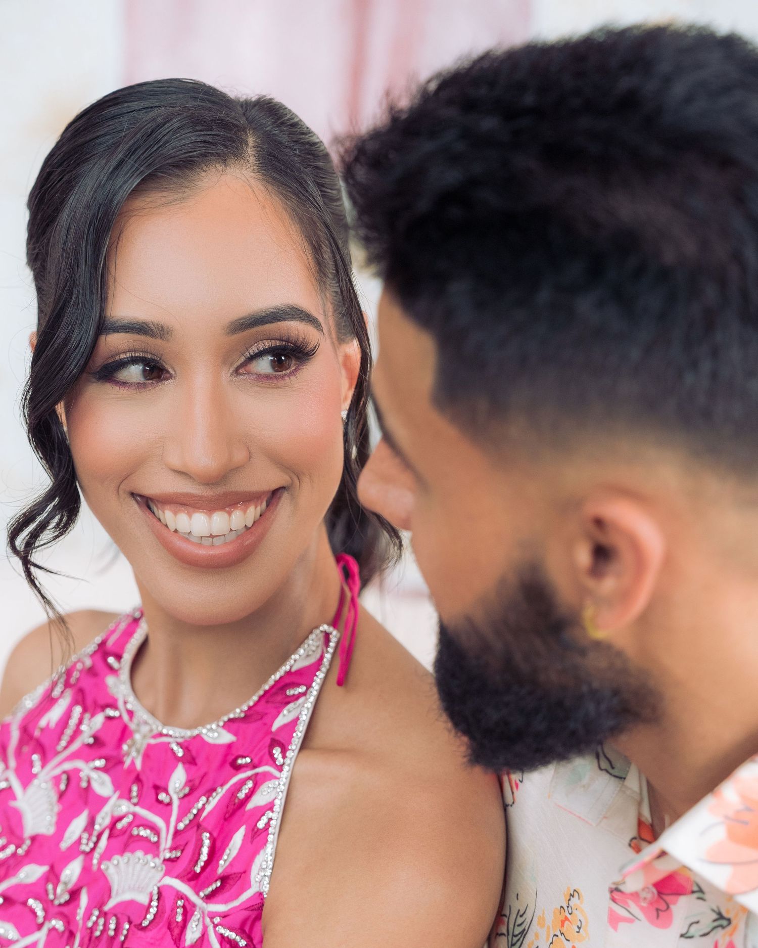Close-up portrait of a smiling woman in a pink embellished halter top with her bearded partner leaning in beside her.