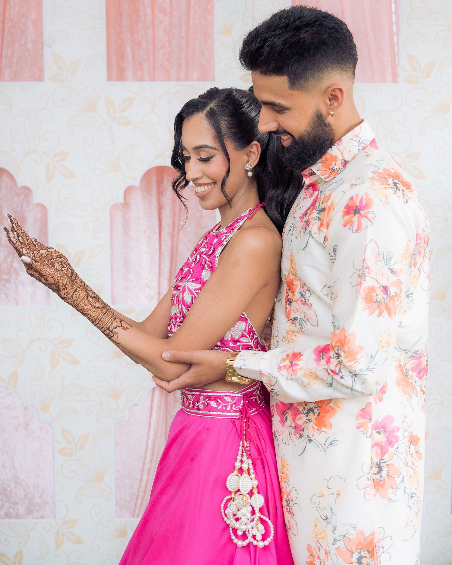 Couple in coordinated pink and floral outfits embrace against a pastel backdrop, her hand adorned with intricate henna designs.