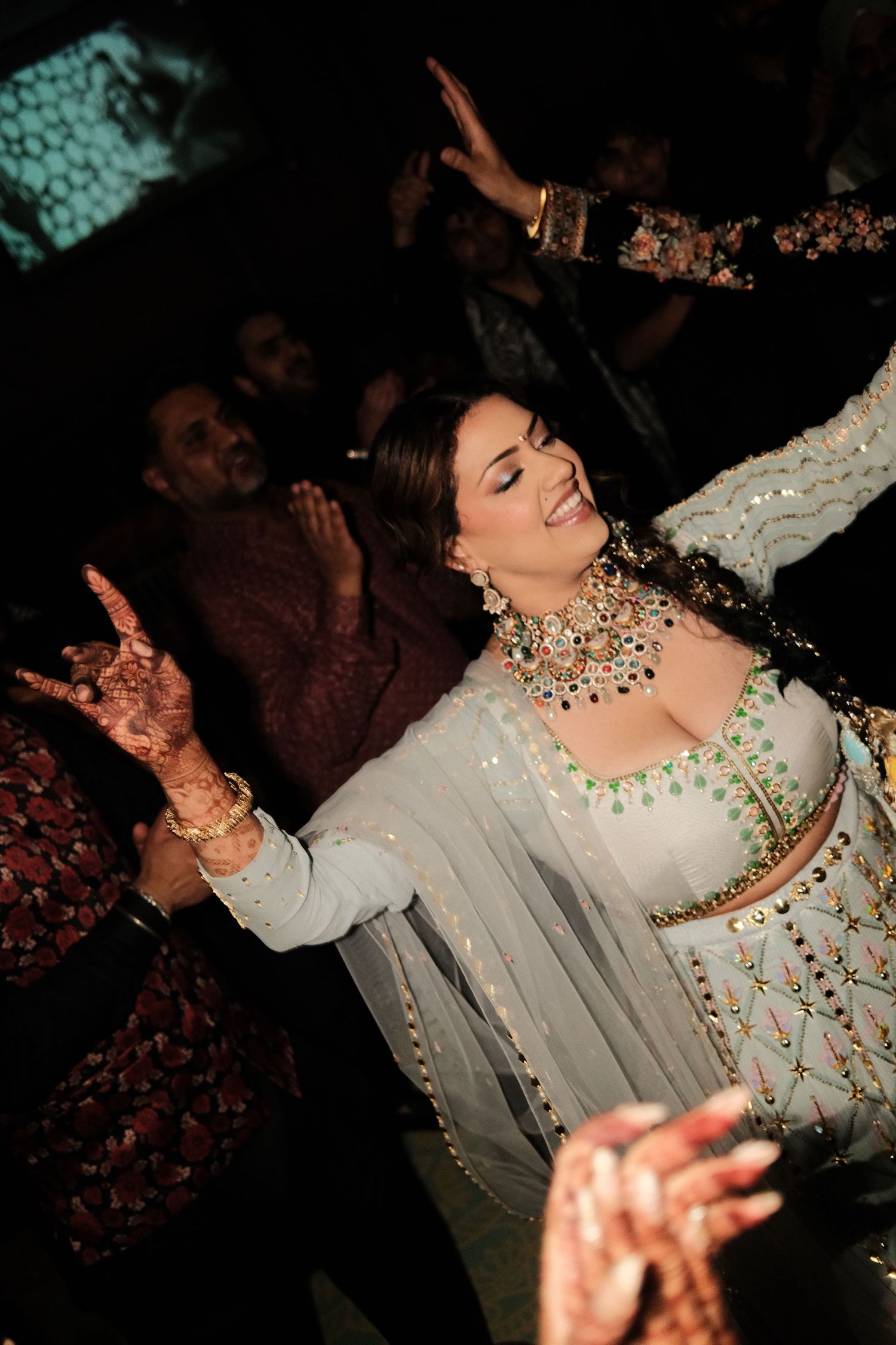 Joyful bride in an embellished mint lehenga and bold jewelry dances with henna-covered hands raised at an Indian wedding celebration.