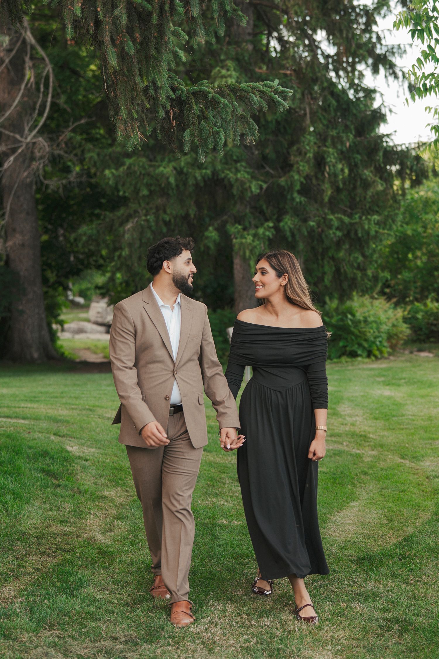 Smiling couple walks hand in hand across a green lawn beneath tall evergreen trees in a candid engagement portrait.