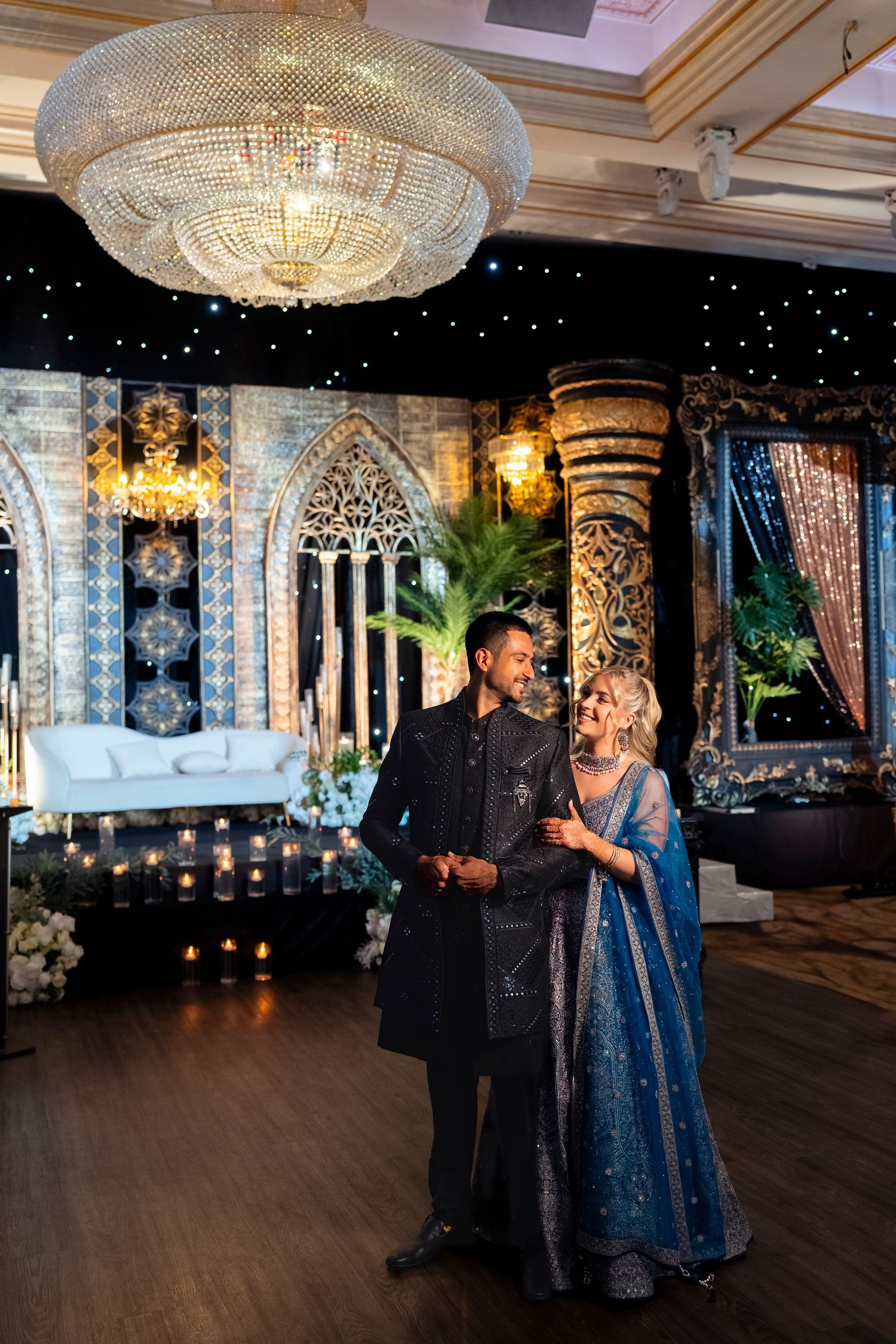 Smiling couple in embroidered black sherwani and blue lehenga pose in a luxurious reception hall with a crystal chandelier and candles.