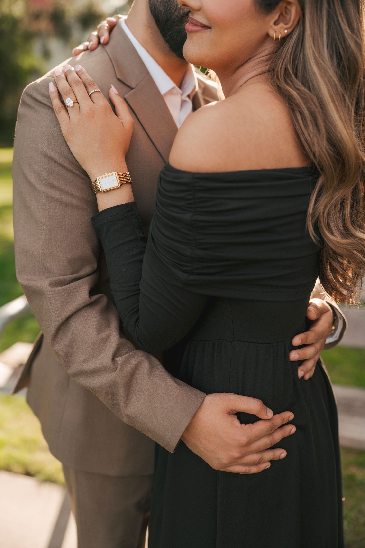 Woman in a black off-the-shoulder dress wearing an engagement ring embraces a man in a tan suit in a romantic outdoor portrait.