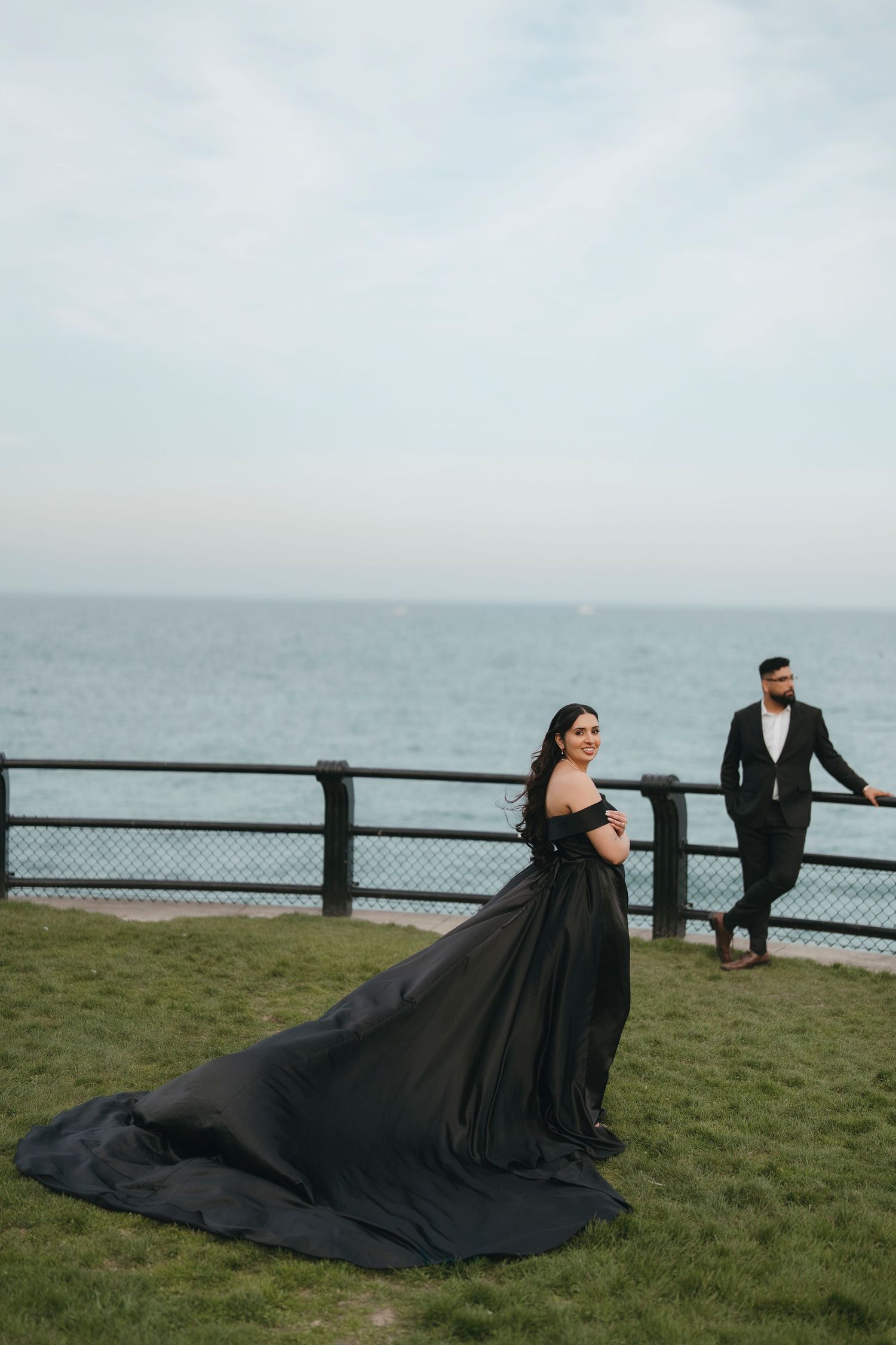 Bride in a black gown with a dramatic train smiles over her shoulder on a grassy lakeside lawn as her fiancé leans on the railing.