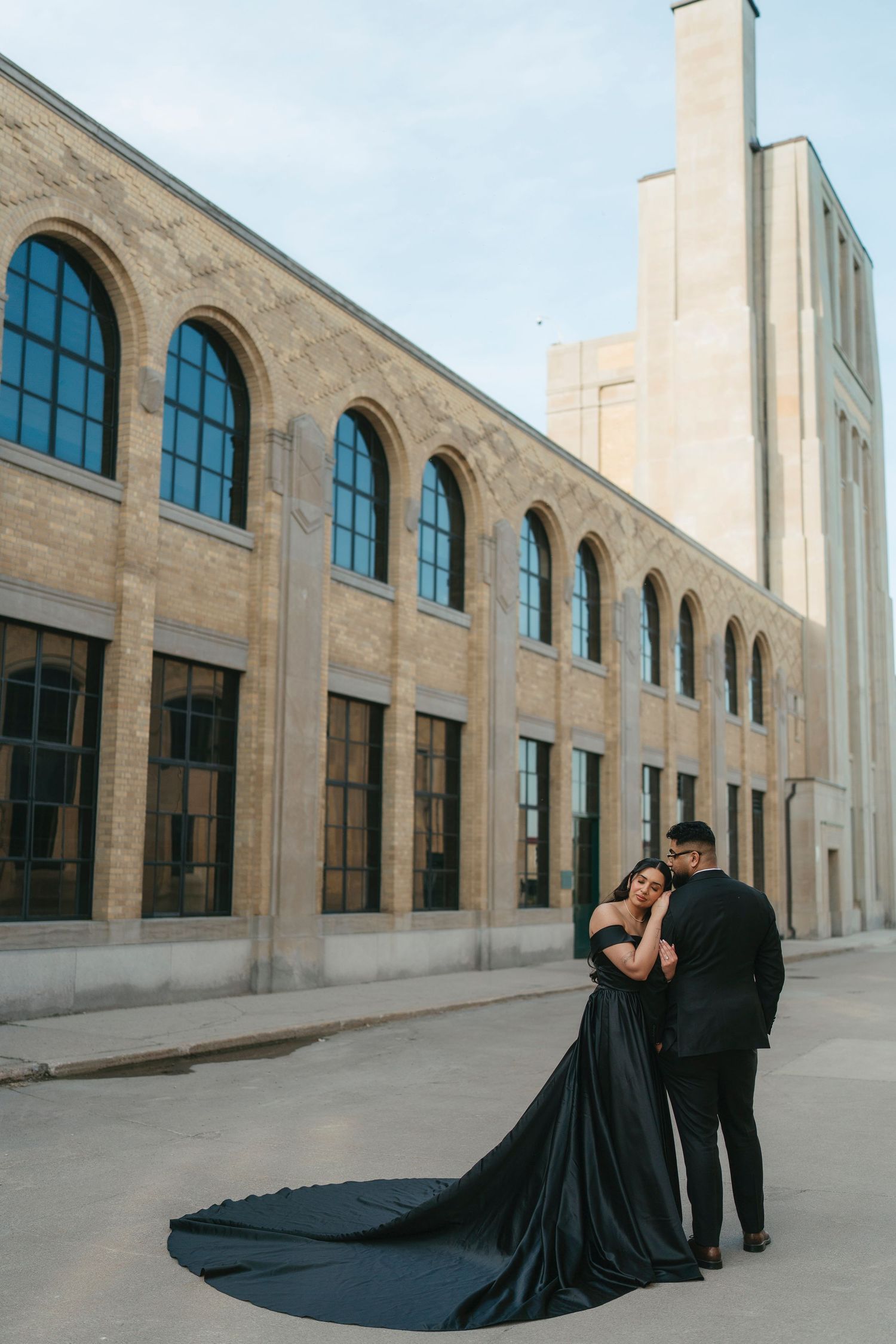 Couple in black formal wear embrace on a quiet street in front of a historic brick building, her long gown train spread behind.