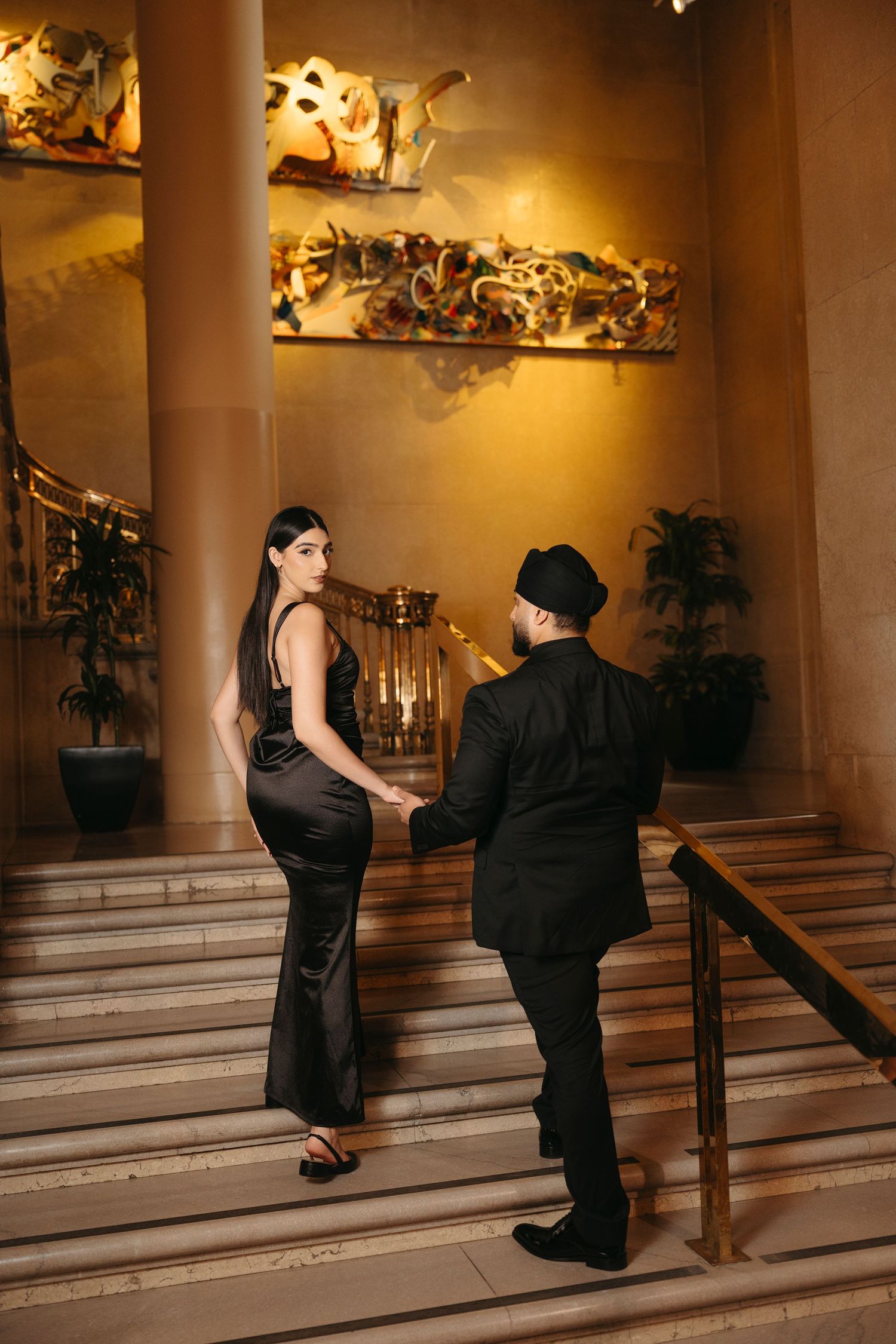 Couple in black outfits hold hands while walking up a stately staircase in a warmly lit elegant lobby.